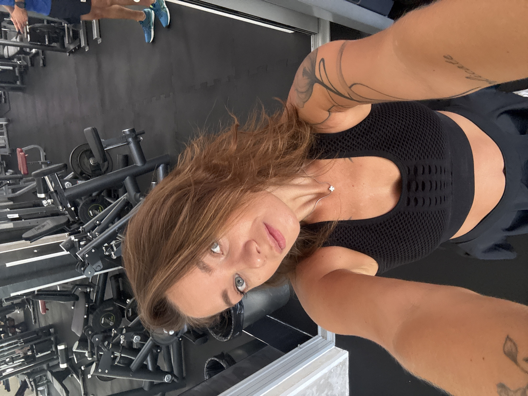 The image shows a woman in a gym taking a selfie. She has long, brown hair and is wearing a black sports bra and black shorts. She has tattoos on her arms. The gym equipment is visible in the background. The lighting suggests it might be daytime. The image has been rotated so the subject is sideways.
