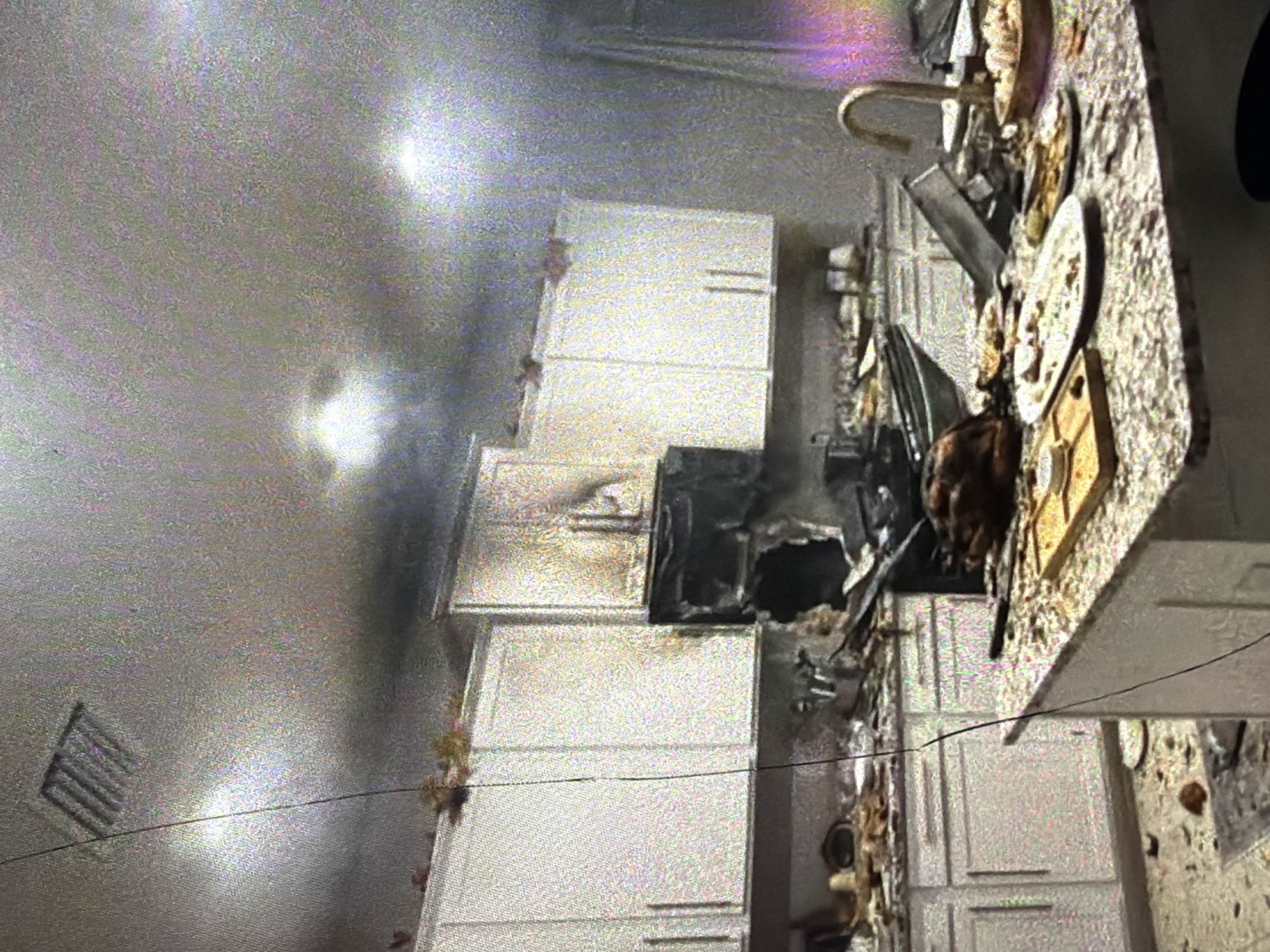 The image depicts the aftermath of a fire in a kitchen. The cabinets above the stovetop are heavily scorched and appear to have sustained significant damage, with a gaping hole in the center of the backsplash area. Debris is scattered across the countertops and floor, including plates with remnants of food, serving dishes, and what appears to be a cooked turkey. A faucet is visible on the countertop. Smoke or soot is present on the ceiling and upper cabinets. The overall scene suggests a substantial fire event that has caused considerable damage to the kitchen. This scene is located in the city of Bukuru, Nigeria.