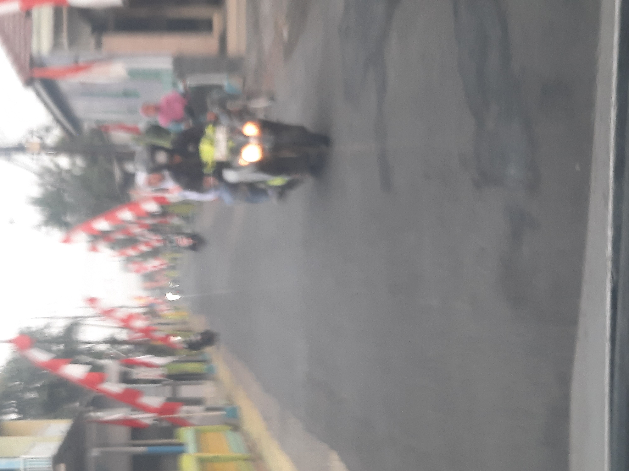 The image depicts a street scene in Banjarkemantren, Indonesia, characterized by decorative red and white flags lining both sides of the road, indicative of a celebration or national holiday. The overall image is blurry and out of focus, making precise identification of details challenging. In the foreground, a motorcycle with its headlights on is visible, carrying at least two individuals. One person, likely the driver, wears a dark helmet. Behind this motorcycle, another person on a separate motorcycle is partially visible. Further down the street, other individuals and motorcycles are present, though their details are obscured by the blurriness and distance. The environment appears to be a residential or commercial area with buildings visible along the sides of the street. The road surface is asphalt. The sky is overcast or hazy, contributing to the soft lighting of the scene.