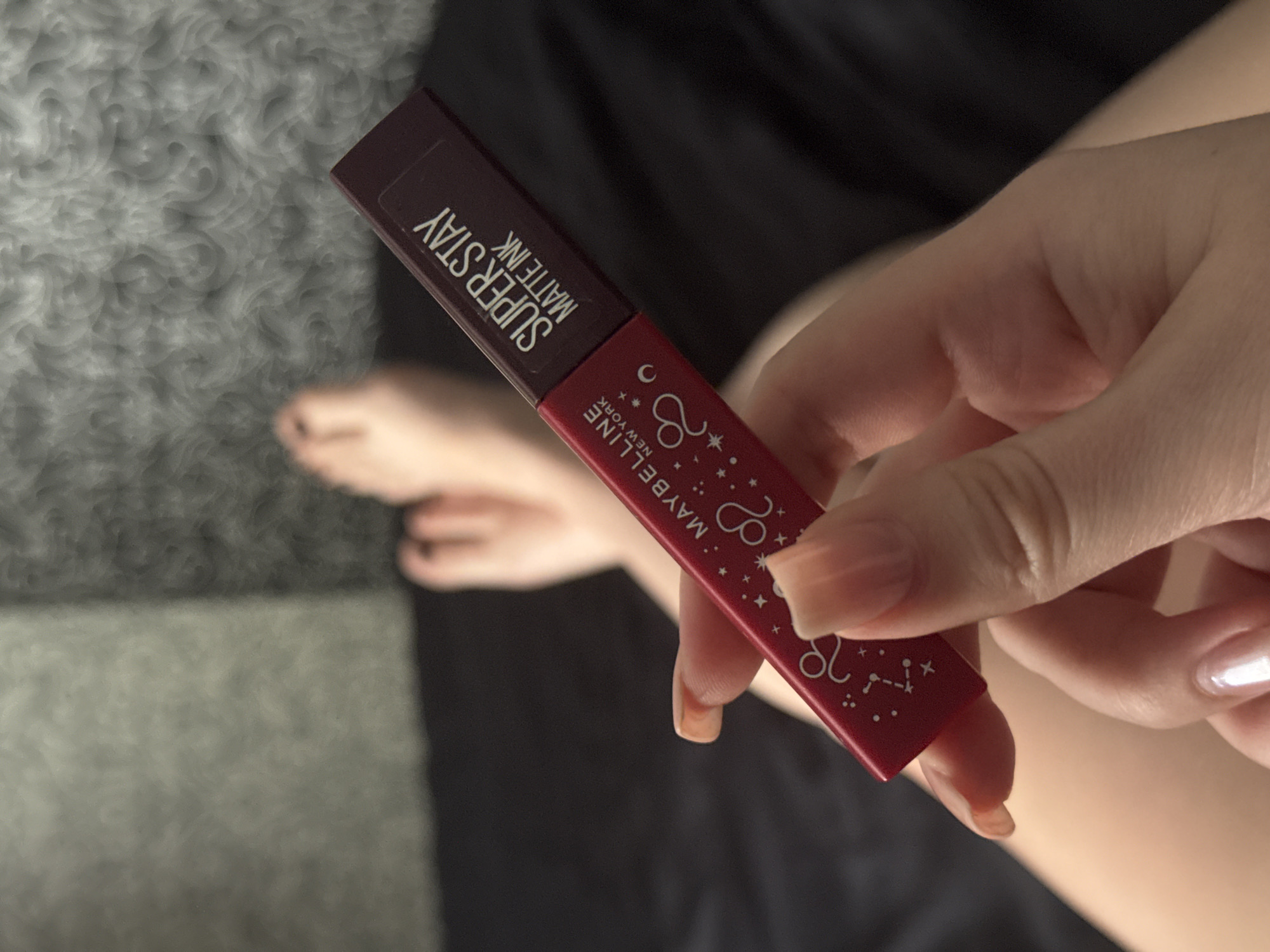 The image shows a close-up of a hand holding a tube of Maybelline Super Stay Matte Ink lipstick. The lipstick tube is red with white celestial graphics, including stars and zodiac signs, and white text that reads "MAYBELLINE NEW YORK" and "SUPER STAY MATTE INK". The hand holding the lipstick has long, manicured nails with a pearlescent finish. In the background, out of focus, are a foot and a dark fabric surface, possibly clothing or bedding. The lighting suggests an indoor setting, possibly during the daytime. The overall mood is casual and personal. The location context is Novosibirsk, Russia.