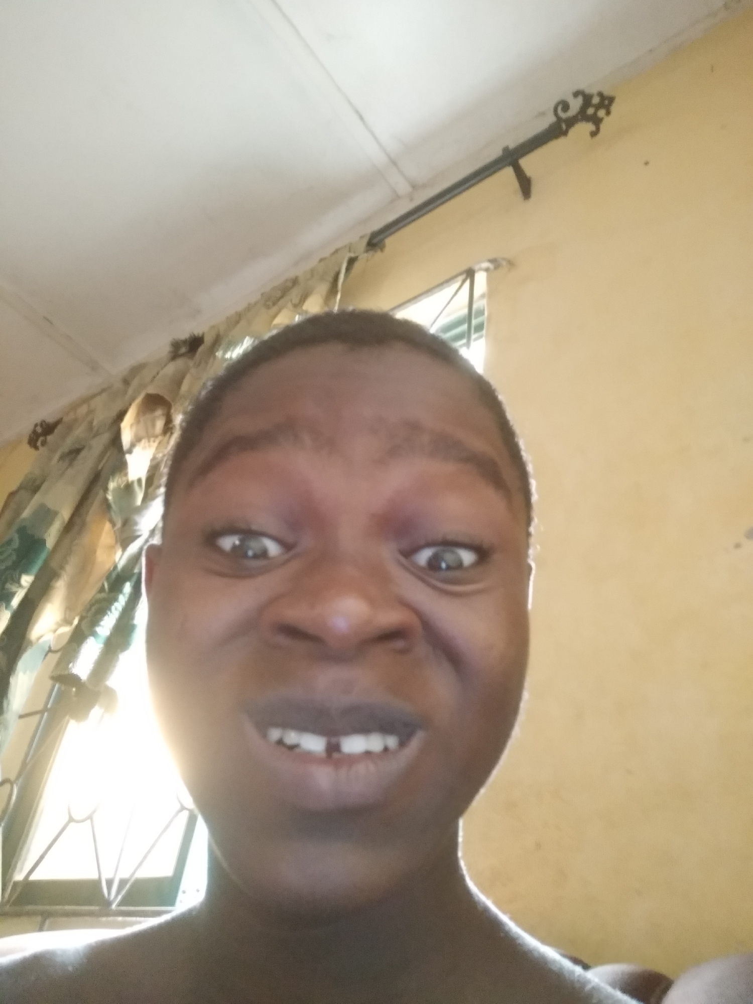 A close-up, slightly low-angle photograph features a young person, likely a child or teenager, with dark skin and short dark hair, posing with an exaggerated, humorous facial expression. Their eyes are wide and slightly bulged, their nose is crinkled, and their mouth is open in a wide smile or grimace, revealing a noticeable gap between their two front teeth. The subject is looking directly at the camera, suggesting a selfie.
The background reveals an indoor setting, likely a room. To the left, patterned curtains, primarily green and cream, are visible and appear gathered. Behind the subject's left side, a window with metal grilles or bars allows bright natural light to stream in, creating a slight halo effect around the subject. On the right side of the frame, a plain light yellow or cream-colored wall is visible, adorned with a black metal curtain rod featuring an ornate, decorative finial. A white ceiling is also seen along the top edge of the image.
The bright lighting indicates it is daytime. The informal setting and the subject's playful expression suggest a casual moment. The location context of Shangisha, Nigeria, implies a domestic interior in that region. No text is visible in the image.
