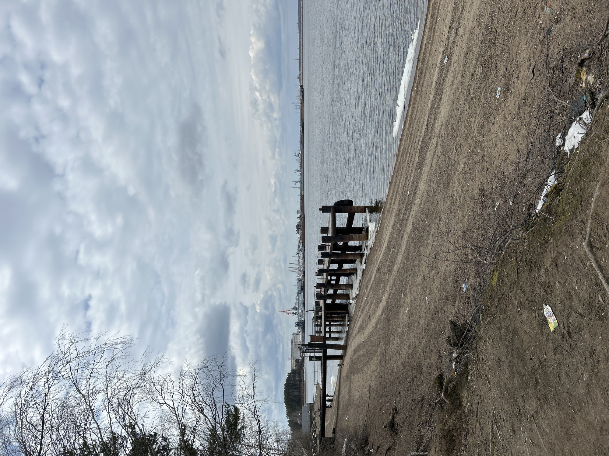The image depicts a waterfront scene in what appears to be the city of Chaikovsky, Russia, on an overcast day. A body of water extends towards the horizon. Along the shoreline, there is a dirt embankment or path. A dilapidated pier or breakwater structure, constructed of dark wood posts and beams, juts out into the water. In the distance, industrial buildings and port infrastructure are visible along the opposite shore, including cranes and other maritime equipment. Bare trees are visible in the foreground. A few scattered patches of snow are near the embankment along with some trash.