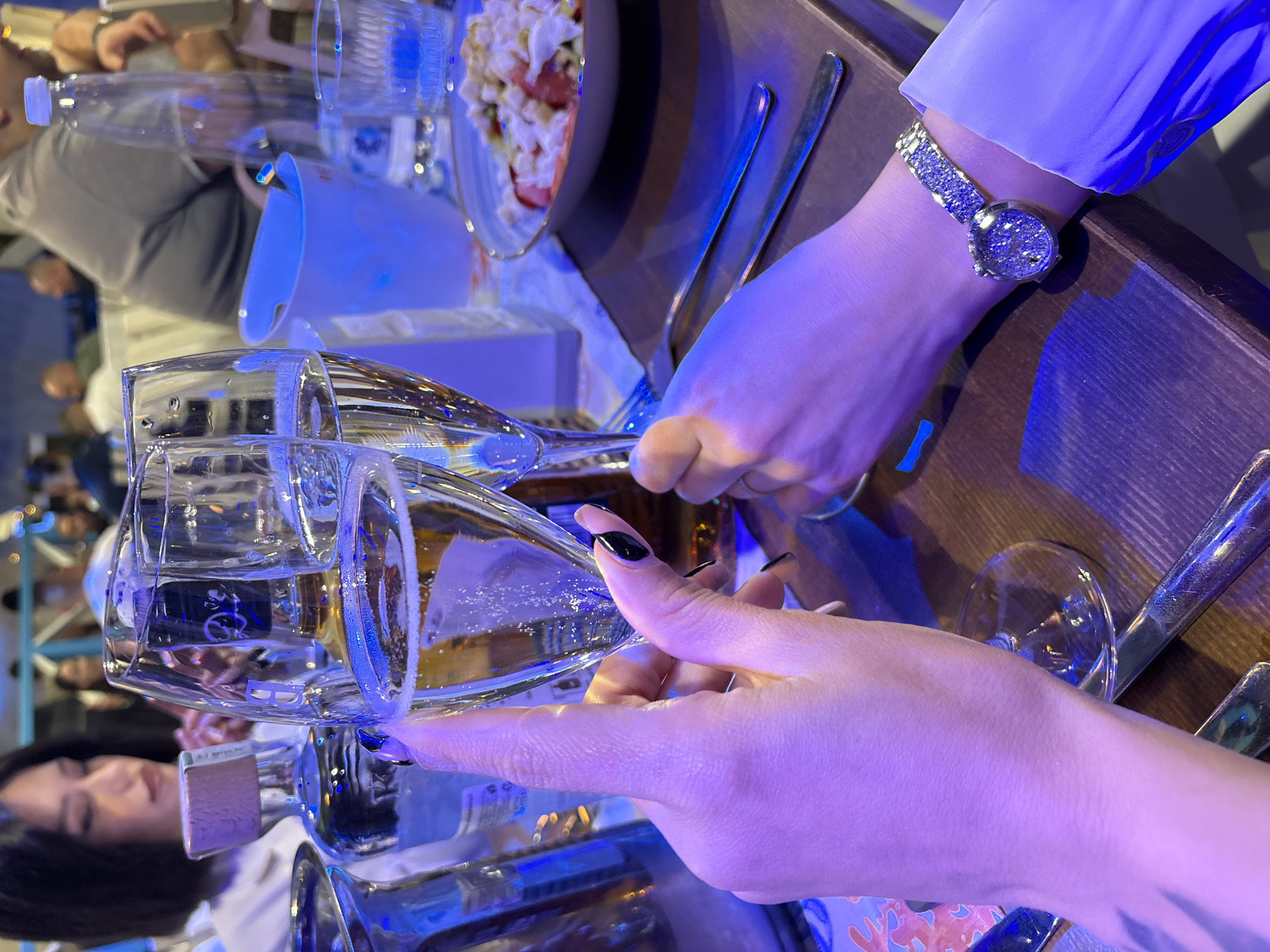 The image shows a dining table setting, likely in an indoor restaurant, although its precise location within Plovdiv, Bulgaria, cannot be determined from visual data. Two champagne flutes are being clinked together. A person wearing a watch with a jeweled band and a light-colored shirt holds one glass. Another person, with black nail polish, holds the other glass. The table also has silverware, a salad, and a water bottle. Other people are visible in the blurred background. The scene is lit with a cool, possibly artificial, light source, giving it a slight blue tint.