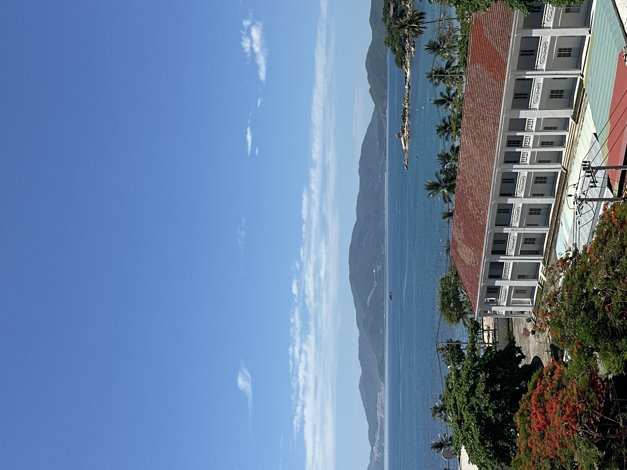 This is a medium-shot, eye-level, vertical photograph of a coastal scene in Nha Trang, Vietnam, under a clear blue sky with scattered white clouds. In the foreground, a long, white building with a red tile roof stretches horizontally across the frame. The building features a colonnaded facade with multiple sets of windows and doors. In front of the building, there are some colorful tarpaulin-covered structures and what appears to be a sports court with a green and red surface.
To the left of the building, lush green trees and a tree with vibrant red flowers are visible. Beyond the building, a wide expanse of calm, blue ocean meets a series of rolling, green-covered mountains in the distance. A small island with rocky outcrops and sparse vegetation can be seen in the middle ground, with a few boats dotting the water around it. Palm trees are scattered along the coastline in the middle ground. The overall impression is one of a tropical resort or coastal town in bright daylight. The specific location within Nha Trang is not identifiable from the visual cues alone, but it depicts a characteristic coastal landscape of the region. There are no people or animals visible in the image.
