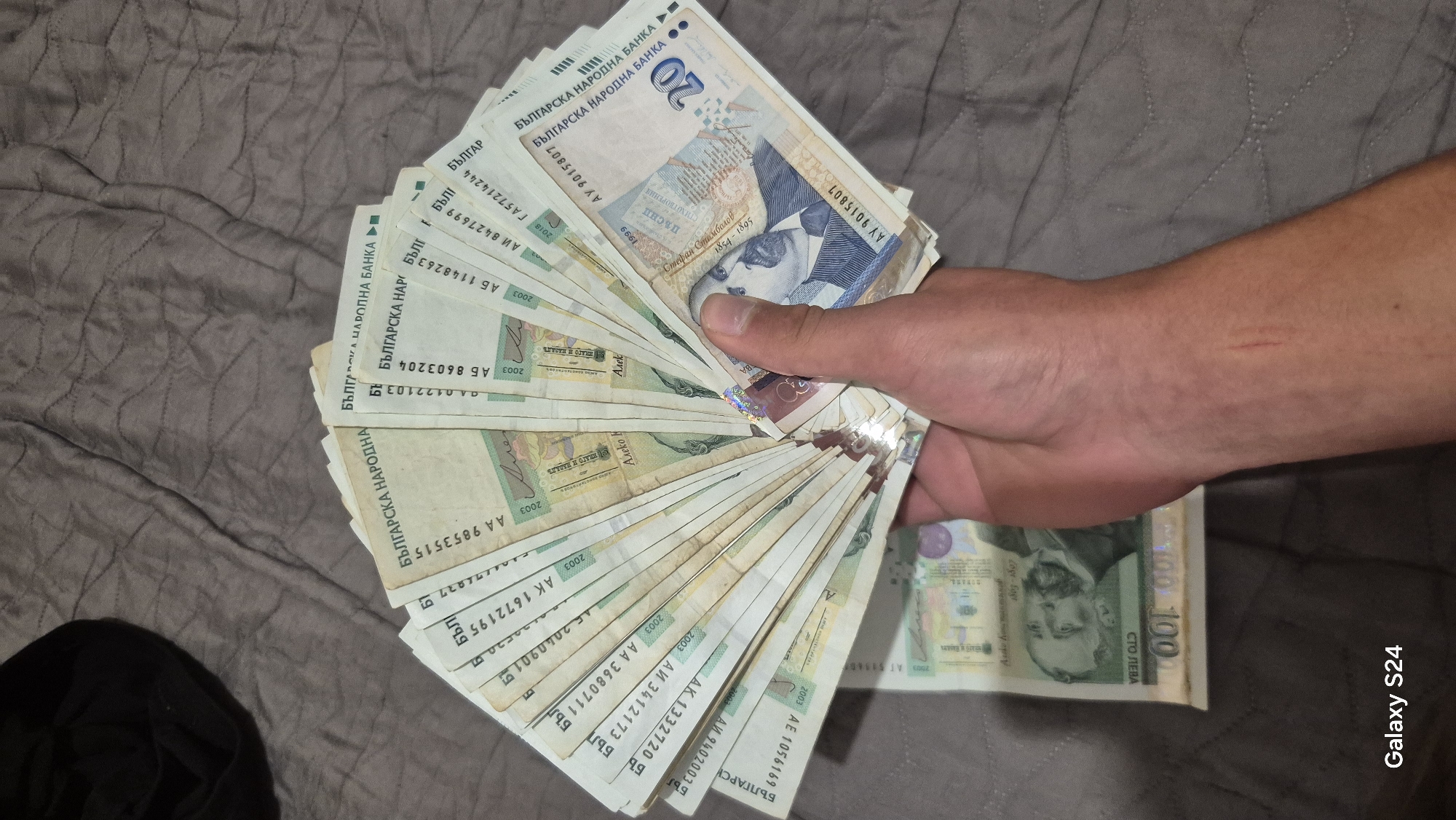 A person's right hand holds and fans out a large stack of Bulgarian banknotes against a textured, grayish-brown fabric background, possibly a blanket or comforter. The hand, with a visible scratch on the forearm, dominates the right side of the frame.
The banknotes are predominantly 20 Bulgarian Lev notes, with one 100 Bulgarian Lev note partially visible at the bottom right. The most prominent 20 Lev note clearly displays the portrait of "Стефан Стамболов" (Stefan Stambolov) with his birth and death years "1854-1895", along with the words "ПЪСНИ" and "СТИХОТВОРЕНИЯ" (Poems). All visible notes feature the text "БЪЛГАРСКА НАРОДНА БАНКА" (Bulgarian National Bank) and various serial numbers. Many notes have an issue year of "2003", while one 20 Lev note shows "2018". Visible serial numbers include AY 9015807, ГА 57214244, АИ 8427699, АБ 1148263, АБ 8603204, АА 9853515, АК 1672195, АИ 34121738, АА 36807118, АК 1332720, АИ 9402003, and АЕ 1056169. A black object, possibly clothing, is visible in the bottom left corner. The image includes a "Galaxy S24" watermark in the bottom right.