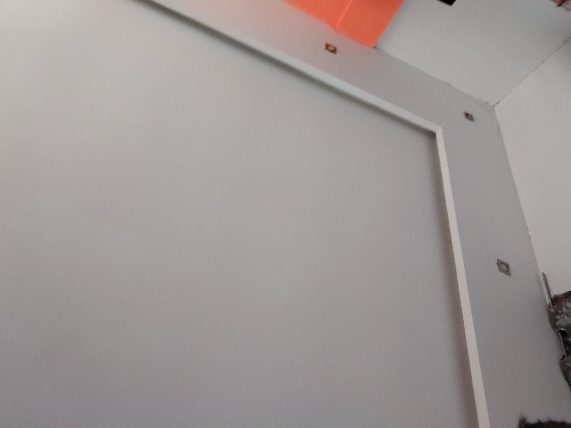 This image captures an interior scene, likely a room or hallway, characterized by minimalist design and neutral colors. The central focus is a large, light grey wall occupying most of the frame. A prominent white rectangular frame or molding is visible on the upper and right sides of this grey wall, suggesting a recessed panel or a decorative architectural element. This frame extends along the top, partially on the right edge, and corners the grey wall.
In the upper-right corner of the image, the perspective shifts to show a corner of the room, where the light grey wall meets an adjacent white wall and a white ceiling. Small, dark, square-shaped indentations or holes are visible on the white walls and within the white frame, possibly indicating mounting points, electrical outlets, or unfinished features. A bright orange object or surface is partially visible along the top edge of the image, contrasting with the dominant grey and white palette.
The lower-right section of the image reveals a cluttered area with dark materials, possibly fabrics, debris, or stored items, leaning against the white wall. This suggests the space might be under construction, renovation, or is a utility corner. The lighting appears uniform, indicative of indoor conditions, and no people or specific activities are depicted. There is no visible text in the image. The overall impression is of a contemporary, somewhat unfinished interior space in Santa Ana, Venezuela.