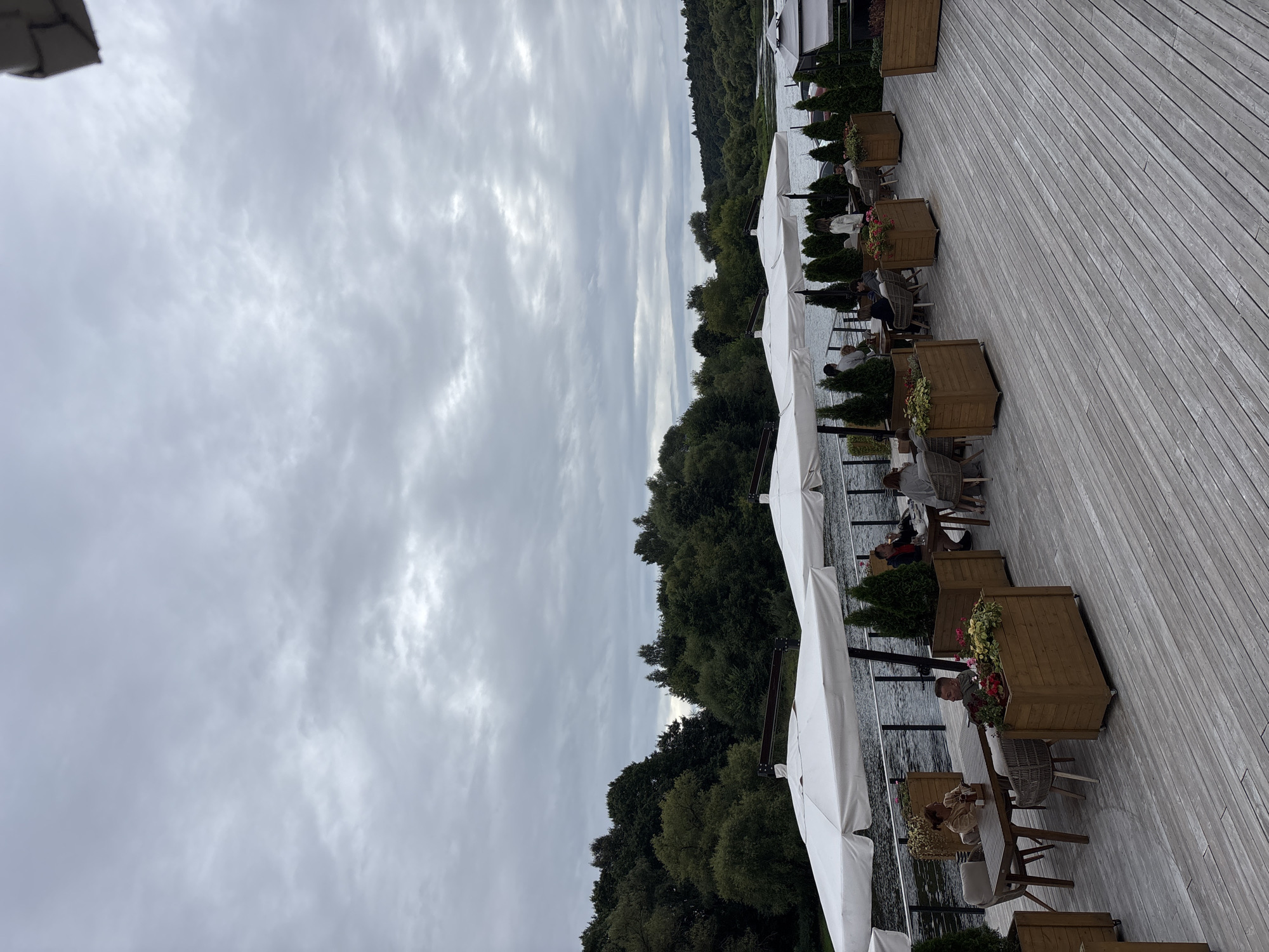 This image depicts an outdoor dining area at a restaurant located on the Moskva River in Zvenigorod, Russia. The scene is set on a wooden deck with several tables and chairs arranged under white umbrellas, suggesting a dining establishment that caters to patrons seeking an outdoor experience by the water.
In the foreground, the wooden deck is visible, laid with planks running diagonally across the image. Several large wooden planters filled with greenery and flowers are strategically placed between tables, adding a touch of natural decoration to the area.
Several patrons are seated at the tables, engaged in dining or conversation. In the middle ground, a woman in a light-colored top is seated at a table with another person, their meal visible. Further down the deck, more individuals are seen enjoying their time outdoors, with some seated at tables and others standing.
The backdrop of the scene features the Moskva River, its surface appearing calm and reflecting the overcast sky. Beyond the river, a dense forest of green trees lines the bank, creating a natural and serene environment. The sky is predominantly cloudy, with a diffused light suggesting a cool or temperate day. The overall atmosphere is one of relaxed outdoor dining, with the natural beauty of the river and forest providing a scenic backdrop.