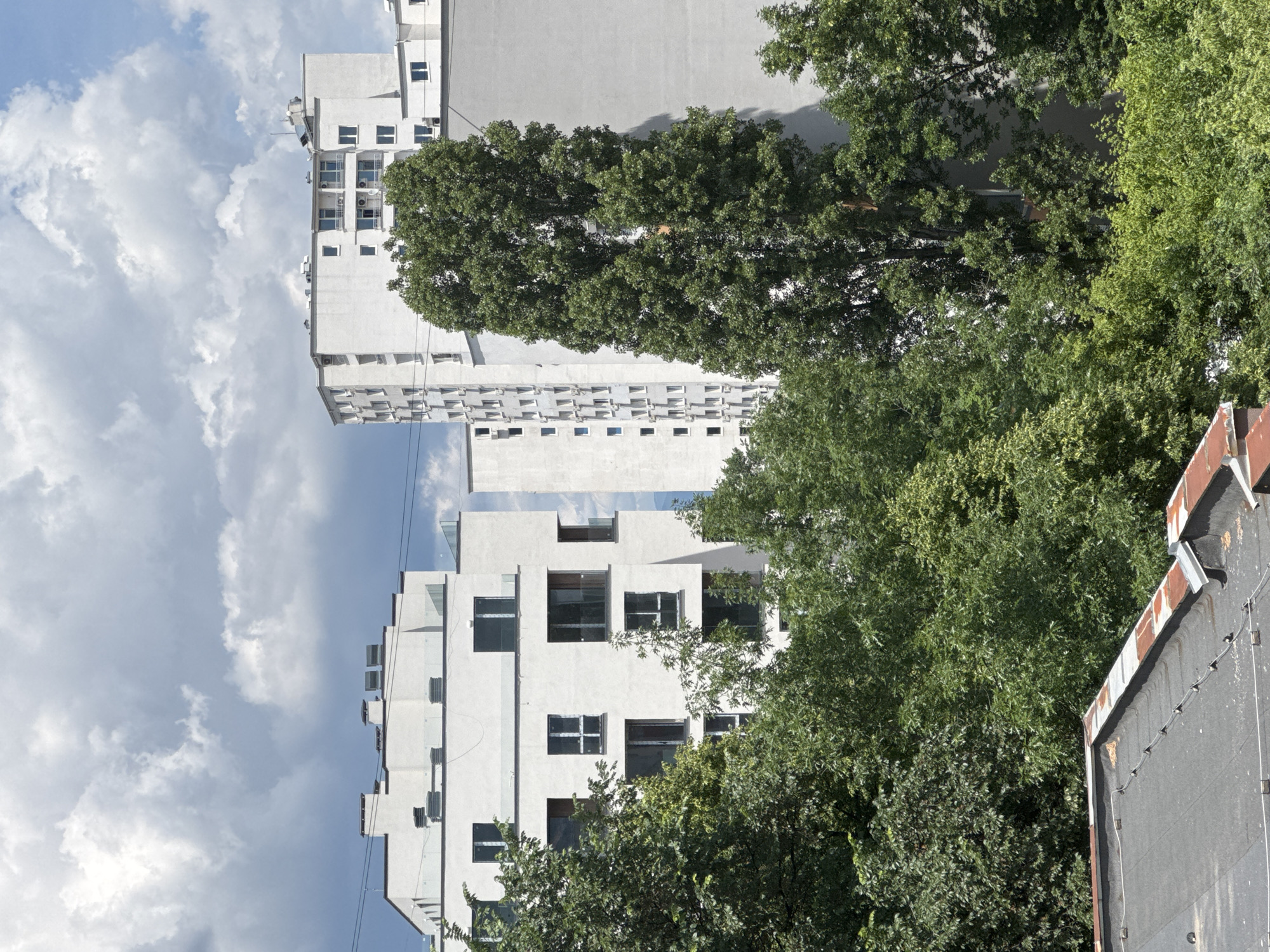 The image depicts an urban scene in Sofia, Bulgaria, characterized by a juxtaposition of modern architecture and lush greenery. In the foreground, a portion of a flat, dark-grey rooftop with rusted metal edging and visible wires suggests an older structure. Behind this, a dense canopy of mature green trees dominates the mid-ground, with various shades of green indicating different tree species and their foliage.
Beyond the trees, several modern white apartment buildings rise into the frame. The closest building on the left is multi-story with a flat roof and numerous uniformly spaced windows. Another taller, more complex building stands in the center, featuring a mix of recessed and projecting sections, balconies, and air conditioning units. To the right, a substantial portion of another large, plain white building is visible. These structures are modern, with clean lines and a consistent white facade.
The sky above is a dynamic mix of white, puffy cumulus clouds against a backdrop of pale blue. The lighting suggests a daytime scene, likely late morning or early afternoon, with shadows cast by the buildings and trees. The overall impression is one of a developing urban area where new construction is integrated with existing mature trees and older rooftops. No people are visible in the image.