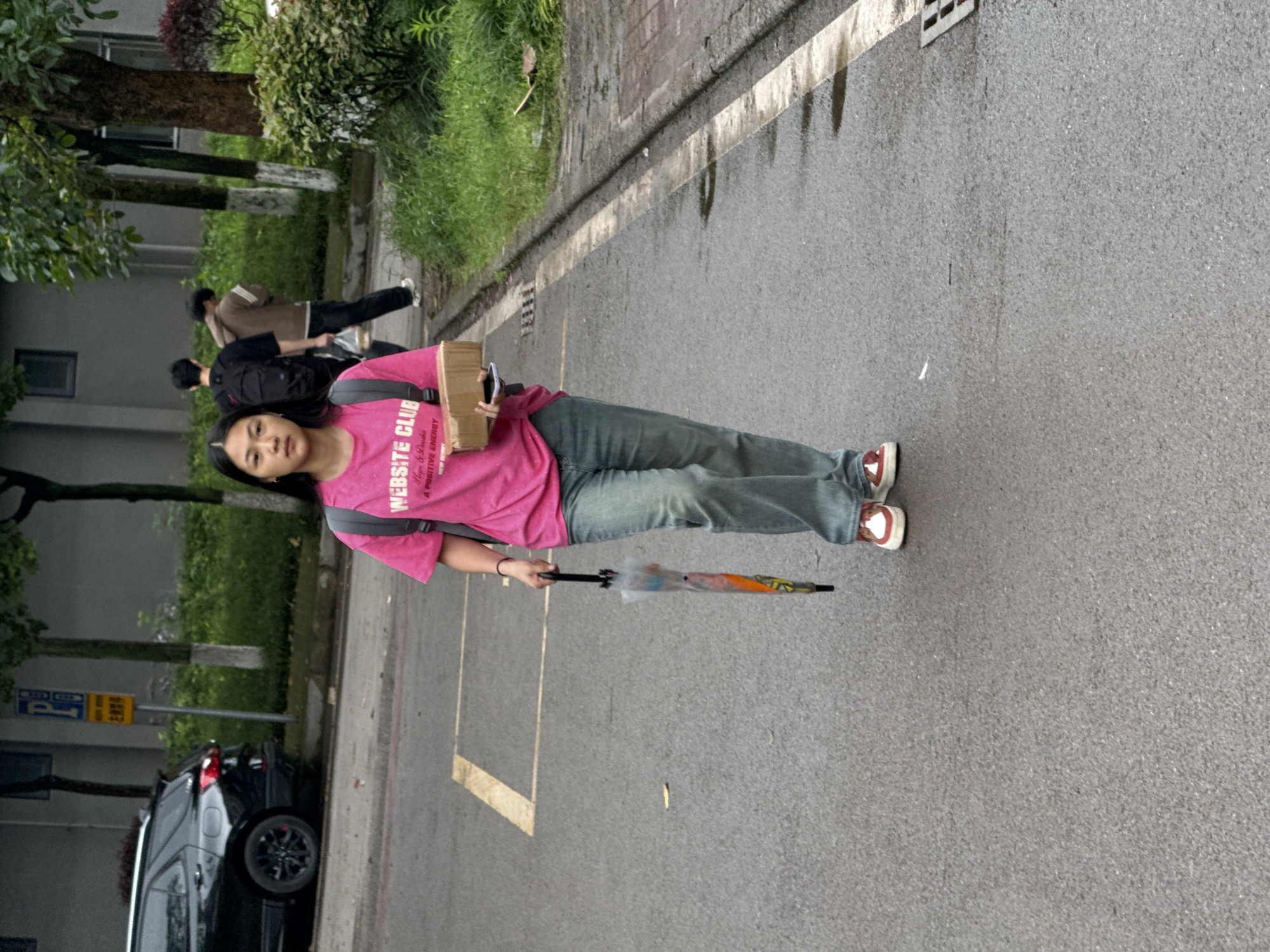 A woman stands on an asphalt road in Jiangjin, China. She wears a pink t-shirt, blue jeans, and red and white sneakers. She is holding a cardboard box in one arm and an umbrella in the other. Two people are walking in the background near a grey building. A black car is parked on the side of the road near a parking sign. Grass and trees are visible in the background.