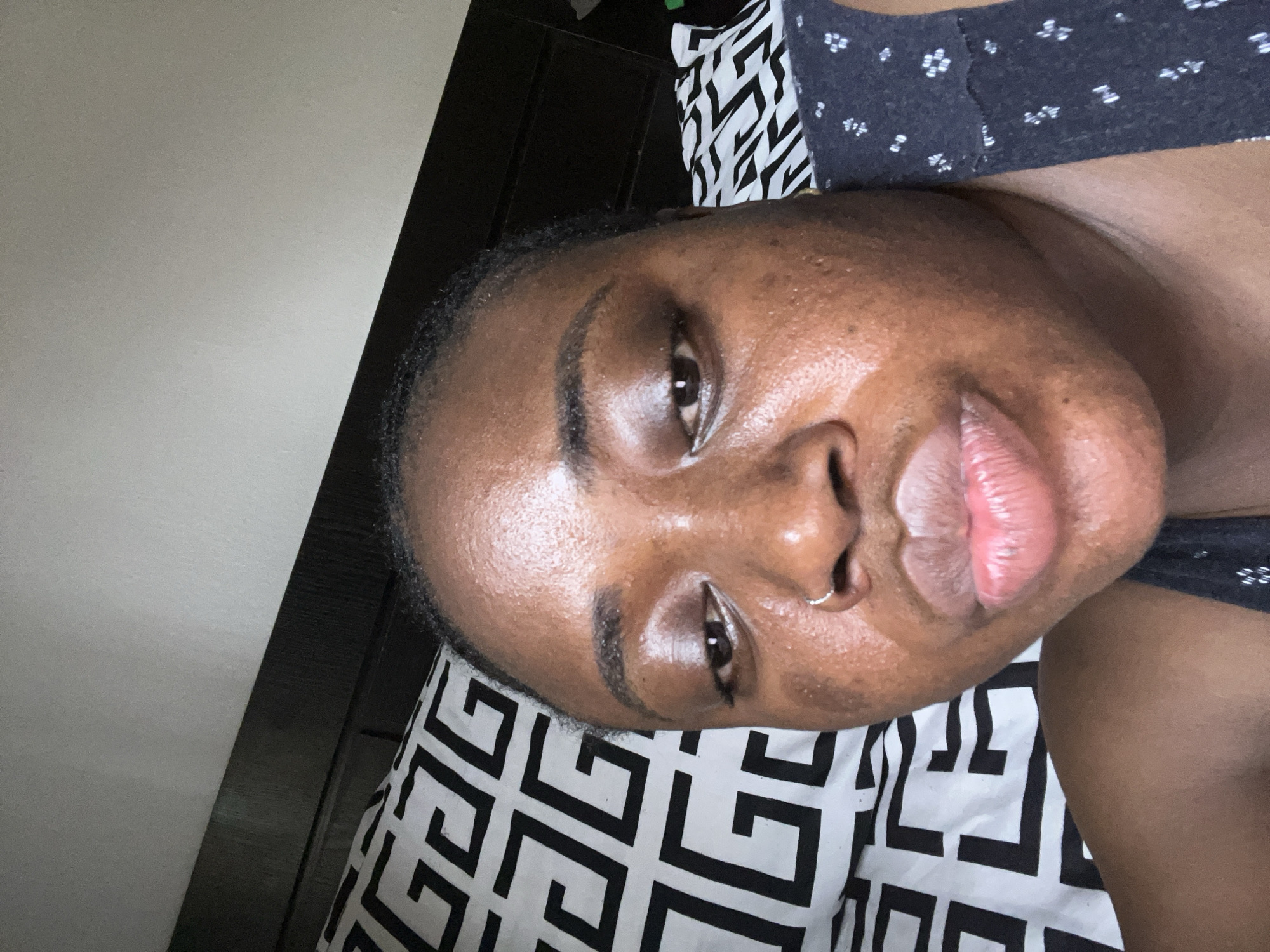 A close-up, rotated portrait of a dark-skinned woman with a silver nose ring is shown. She has dark eyebrows and eyes. Her lips are full and pink. The background includes a black and white patterned fabric and a dark wooden panel. A plain beige wall is also visible. The location is not discernible from the image.