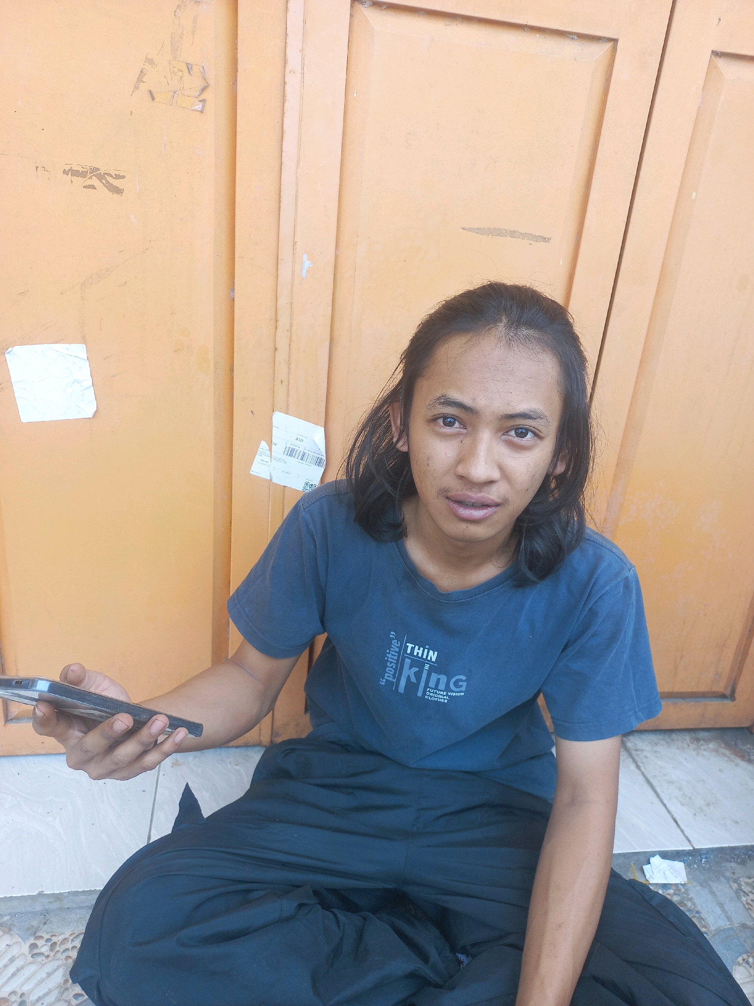 A young man with long, dark hair and a blue t-shirt is sitting cross-legged on the ground in front of an orange door. He is holding a smartphone in his left hand. The t-shirt has text that reads "THIN king POSITIVE". He appears to be looking at the camera. The environment suggests an outdoor urban setting in Baturetno, Indonesia, with a tiled ground and the textured surface of the orange door. There are a few pieces of paper or stickers on the door.