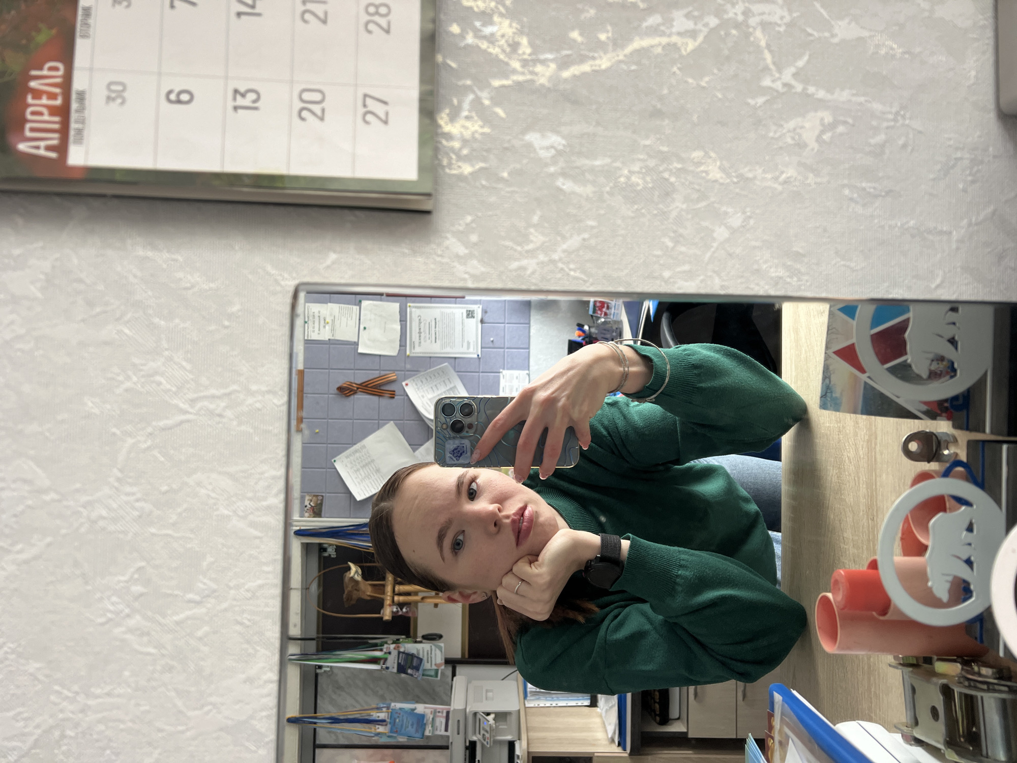 A woman wearing a green sweater is taking a selfie in a mirror. The reflection in the mirror reveals a desk with various objects, including orange cylinders with a white animal print sticker, books, and stationery. Behind the desk is a tiled wall with papers and documents attached to it. A calendar showing the month of April and the numbers 6, 13, 20, 27, and 30 is visible at the top of the frame. The calendar header "АПРЕЛЬ" indicates the month is April in Russian. Based on the objects and text in the image, the location is likely in an office or workspace in Chaikovsky, Russia.