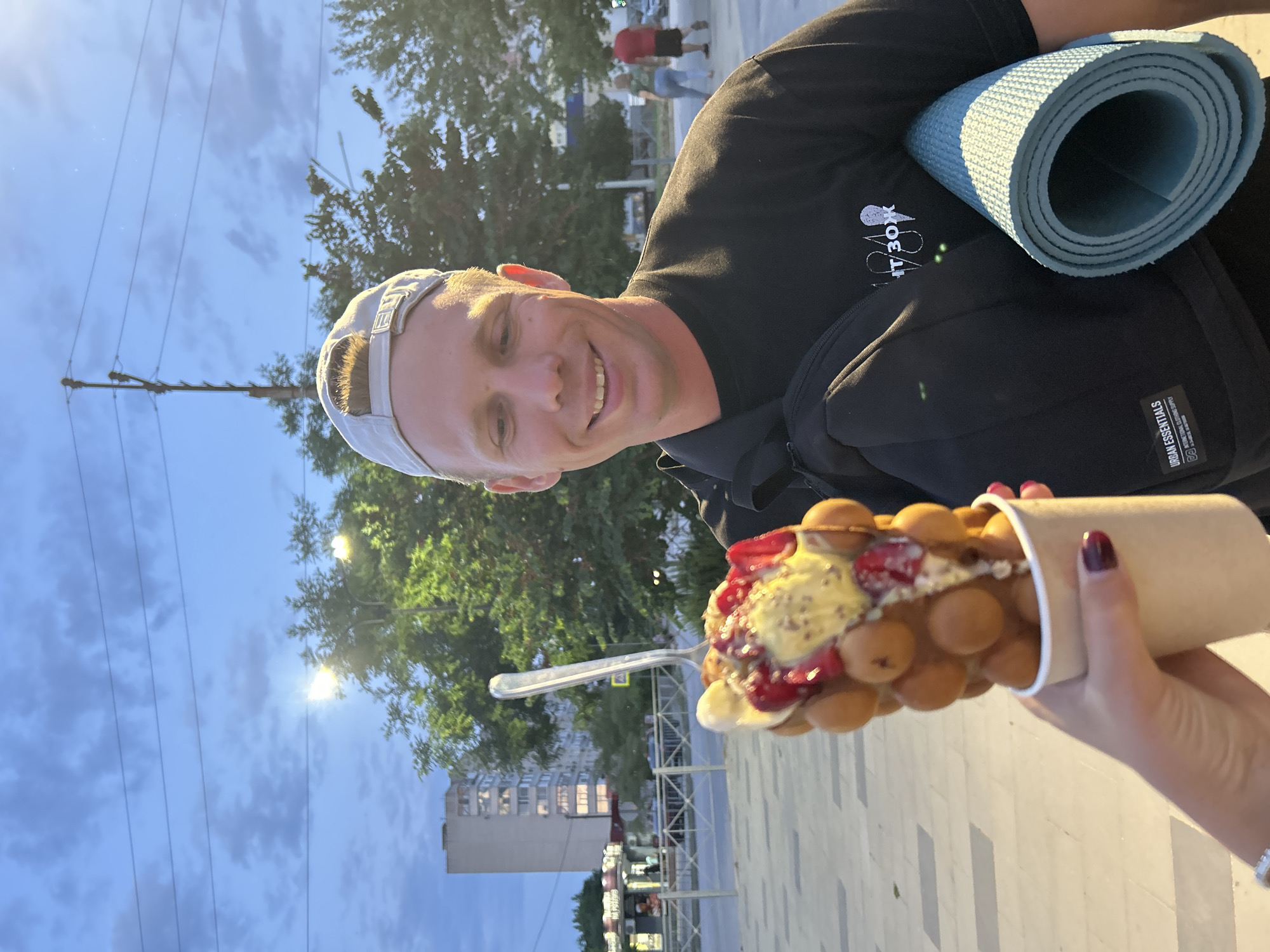 A person wearing a black t-shirt and a grey baseball cap is smiling at the camera. They are holding a bubble waffle cone filled with ice cream, strawberries, and a sauce, with a spoon in it. A rolled-up blue yoga mat is visible on their shoulder, along with a black bag. The background shows trees, a building, streetlights, and power lines against a cloudy sky. The lighting suggests it is either early morning or late afternoon. The image appears to be taken in an urban outdoor setting. There is visible text on the bag that reads "URBAN ESSENTIALS" and a logo that is difficult to discern.