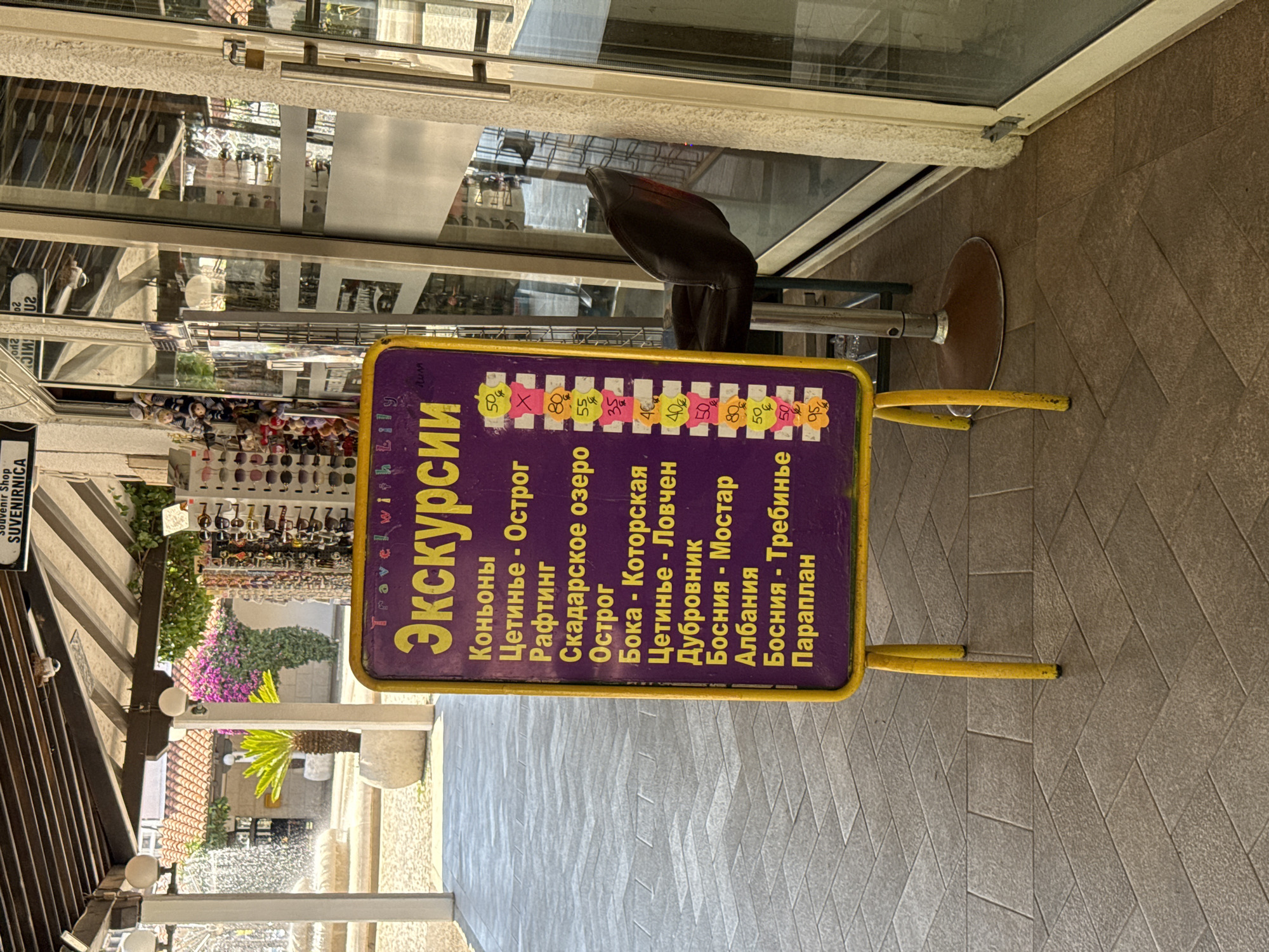 The image depicts a street-level view of a souvenir shop in Budva, Montenegro. A prominent purple and yellow A-frame sign advertising excursions is positioned in front of the shop's entrance. The sign features a list of excursions in Cyrillic script, with prices indicated in Euros (or a similar currency symbol). Excursions listed include "Коньоны" (Conies), "Цетинье - Острог" (Cetinje - Ostrog), "Рафтинг" (Rafting), "Скадарское озеро" (Skadar Lake), "Острог" (Ostrog), "Бока - Которская" (Boka - Kotor), "Цетинье - Ловчен" (Cetinje - Lovcen), "Дубровник" (Dubrovnik), "Босния - Мостар" (Bosnia - Mostar), "Албания" (Albania), "Босния - Требинье" (Bosnia - Trebinje), and "Параплан" (Paragliding). Prices range from 35 to 95.
The shop's facade is visible, with large glass doors and windows reflecting the surroundings. A dark leather bar stool is placed just inside the entrance. To the left of the sign, a fountain and lush greenery suggest a public square or promenade. Various souvenirs, including sunglasses and plush toys, are displayed on racks inside the shop, visible through the glass. The ground is paved with interlocking tiles arranged in a herringbone pattern. A sign above the shop entrance reads "Souvenir Shop" and "SUVENIRNICA" in Cyrillic. The overall scene is bright and sunny, suggesting daytime. No people are clearly visible in the immediate vicinity of the sign or shop entrance.