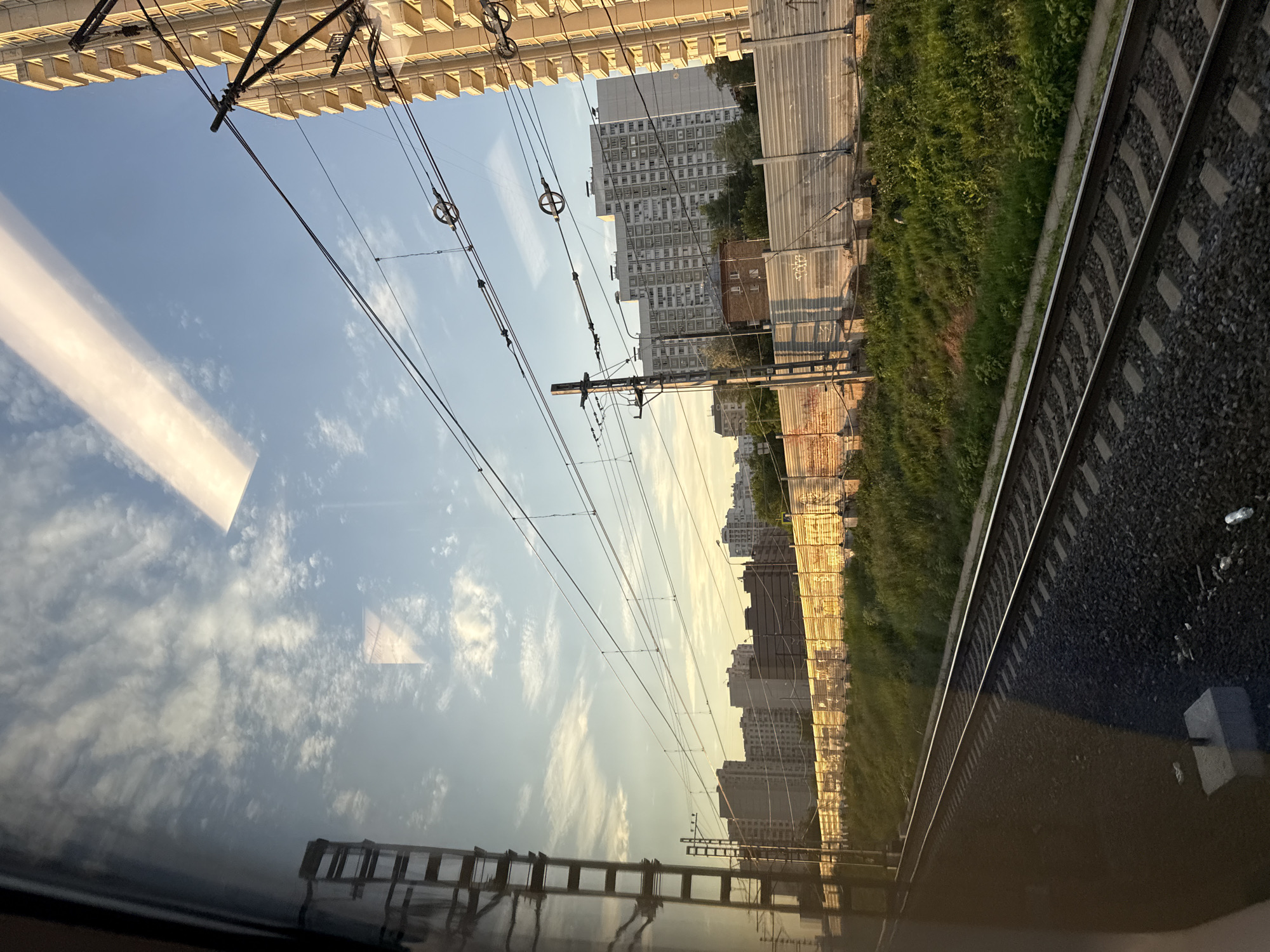 The image captures a view from a moving train, looking out a window. The scene is urban, with a focus on residential buildings and railway infrastructure under a bright, cloud-dotted sky. In the foreground, train tracks are visible, suggesting the observer is on a journey. Along the tracks, there is a grassy embankment and a corrugated metal fence, some parts of which are illuminated by the setting sun, casting a warm glow and revealing graffiti.
Beyond the fence, a series of multi-story apartment buildings line the horizon. Some of these buildings are tall and modern, featuring balconies, while others appear older and more uniform in design. Overhead, a network of electrical wires and support poles are strung across the sky, typical of a railway system. The reflection of a tall, modern building is also visible on the train window, superimposed over the sky and distant cityscape. A long, horizontal cloud formation, bright white and streaked, dominates a section of the blue sky. The overall lighting suggests it is late afternoon or early evening.