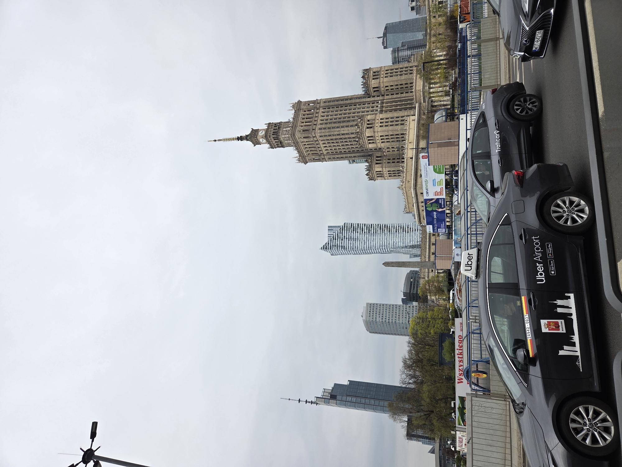 The image captures a view of Warsaw, Poland, dominated by the Palace of Culture and Science in the background. Several modern skyscrapers are visible further back. In the foreground, a row of cars is parked, including an Uber Airport car prominently displaying the company's logo and silhouette of Warsaw skyline. A Trafficar car is visible as well. Street advertisements and signs can also be seen, along with a section of a bridge or pedestrian walkway. The weather appears overcast.