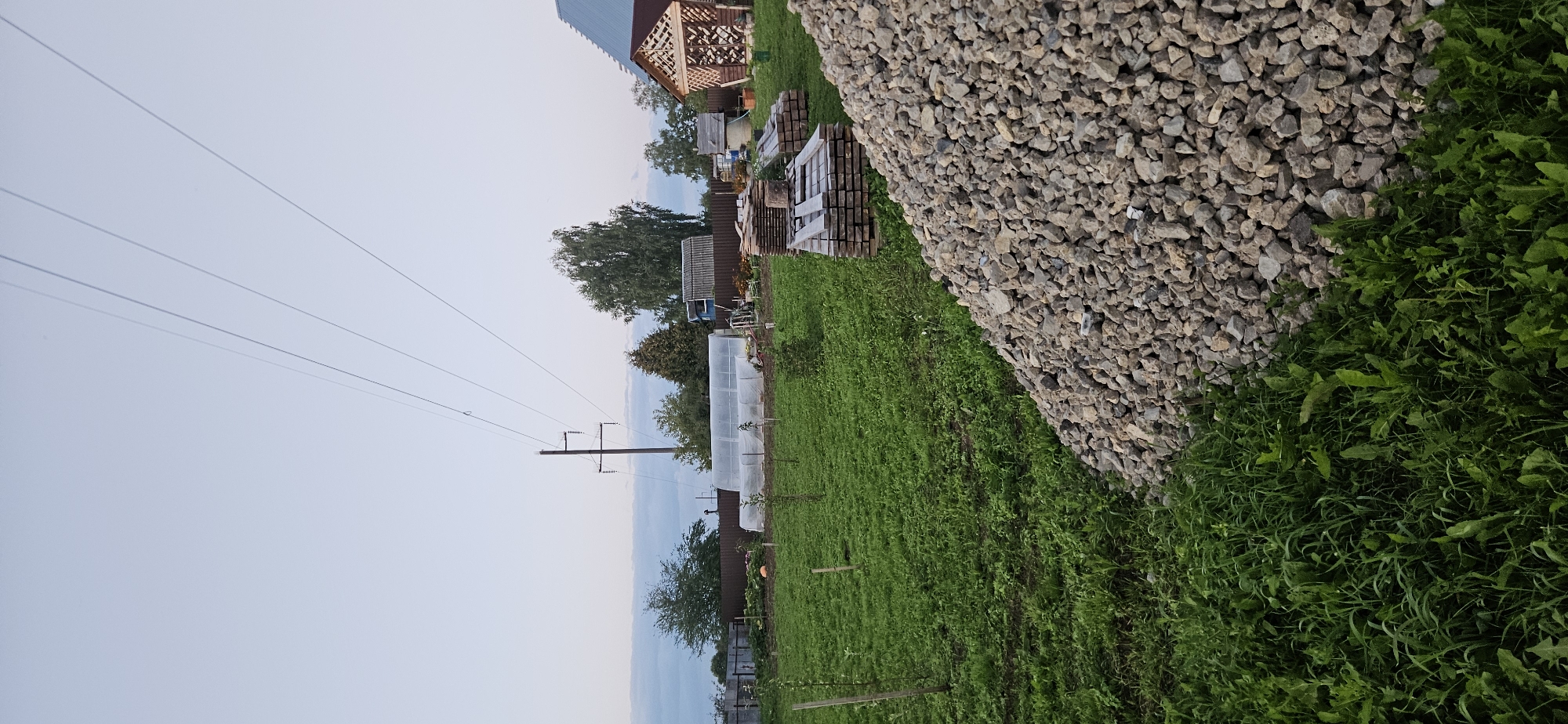 The image depicts an outdoor scene in what appears to be a rural residential area, likely within the Mazalawski Sel'ski Savet in Belarus, judging by the context. In the foreground, a large pile of light-colored gravel dominates the right side of the frame. To the left of the gravel pile, a dense patch of green grass and vegetation grows.
Further into the scene, a well-maintained lawn stretches across the middle ground, dotted with several young trees or saplings. In the background, there are various structures: a greenhouse with a white, translucent covering, a wooden utility pole with power lines extending across the sky, and several other buildings including what looks like a small wooden gazebo with an open lattice structure, and a larger building with a gabled roof. The sky above is a pale, overcast blue, with some faint horizontal cloud formations visible in the distance. The overall impression is one of a developing or actively used residential plot, with construction materials present and evidence of cultivation. There are no people or animals visible in the image.