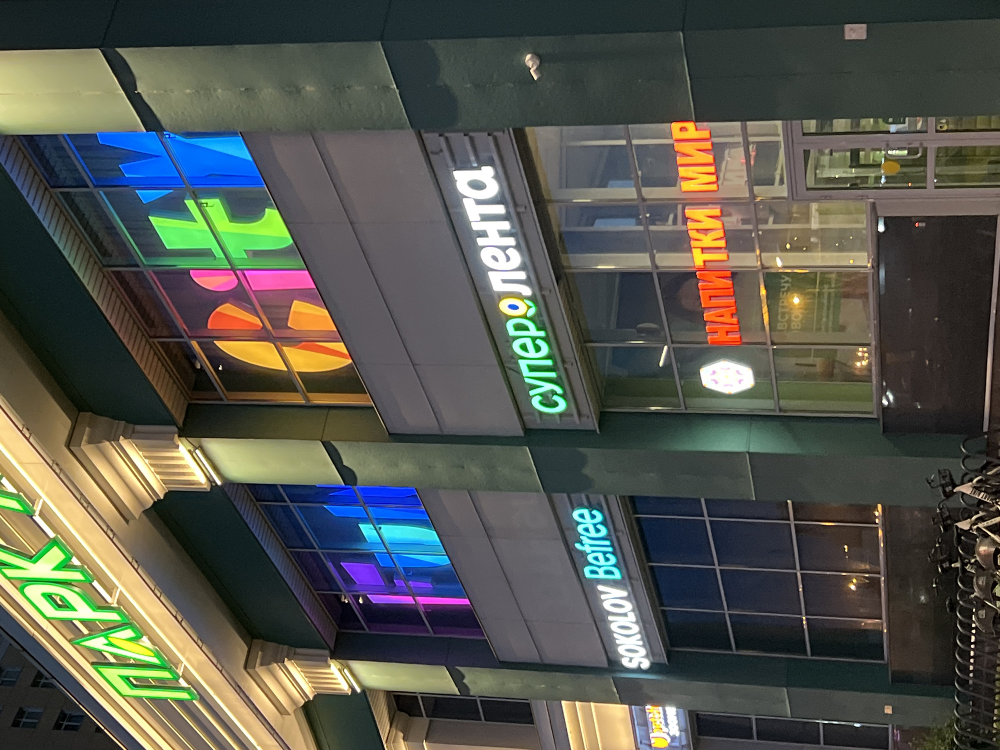 This is a photograph of the exterior of a shopping center at night in Sinyavino, Russia. The building features a dark green facade with large windows. Above the windows, there are illuminated signs for various shops. One prominent sign reads "Super Lenta" in white letters with a colorful, abstract design inside the "o." Another sign in bright orange letters spells out "НАПИТКИ МИР" which translates to "DRINKS WORLD." Below this, a sign for "SOKOLOV Befree" is visible in white letters.
The windows reflect the colorful lights from inside, showing geometric shapes in shades of blue, purple, pink, and yellow. The overall lighting suggests it is nighttime. The scene is urban, likely a commercial district. There are no people visible in the image. The building appears modern. Part of a sign with green neon letters spelling "ПАРК" (PARK) is visible on the left side of the image, angled upwards. The image is slightly tilted, giving a dynamic perspective.