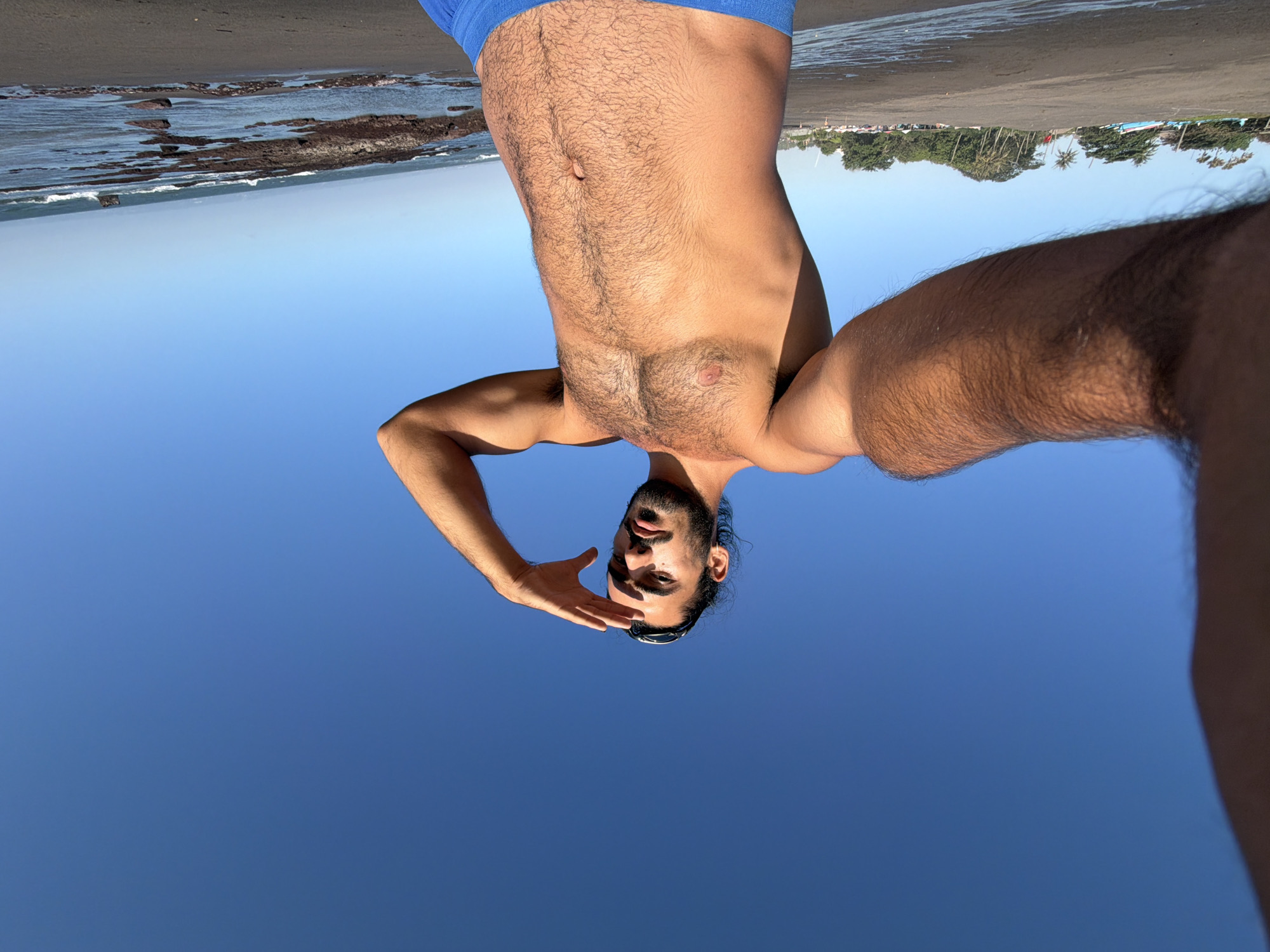 A shirtless man with dark hair, a beard, and sunglasses pushed up on his forehead stands on a sandy beach, his right arm outstretched towards the camera as if taking a selfie. He is wearing blue swim trunks and is shielding his eyes with his left hand, looking towards the horizon. The ocean with waves breaking on rocky shorelines is visible behind him. The background shows a clear blue sky, a line of palm trees, and some small boats on the beach. The location is the beach in Mengwi, Indonesia.