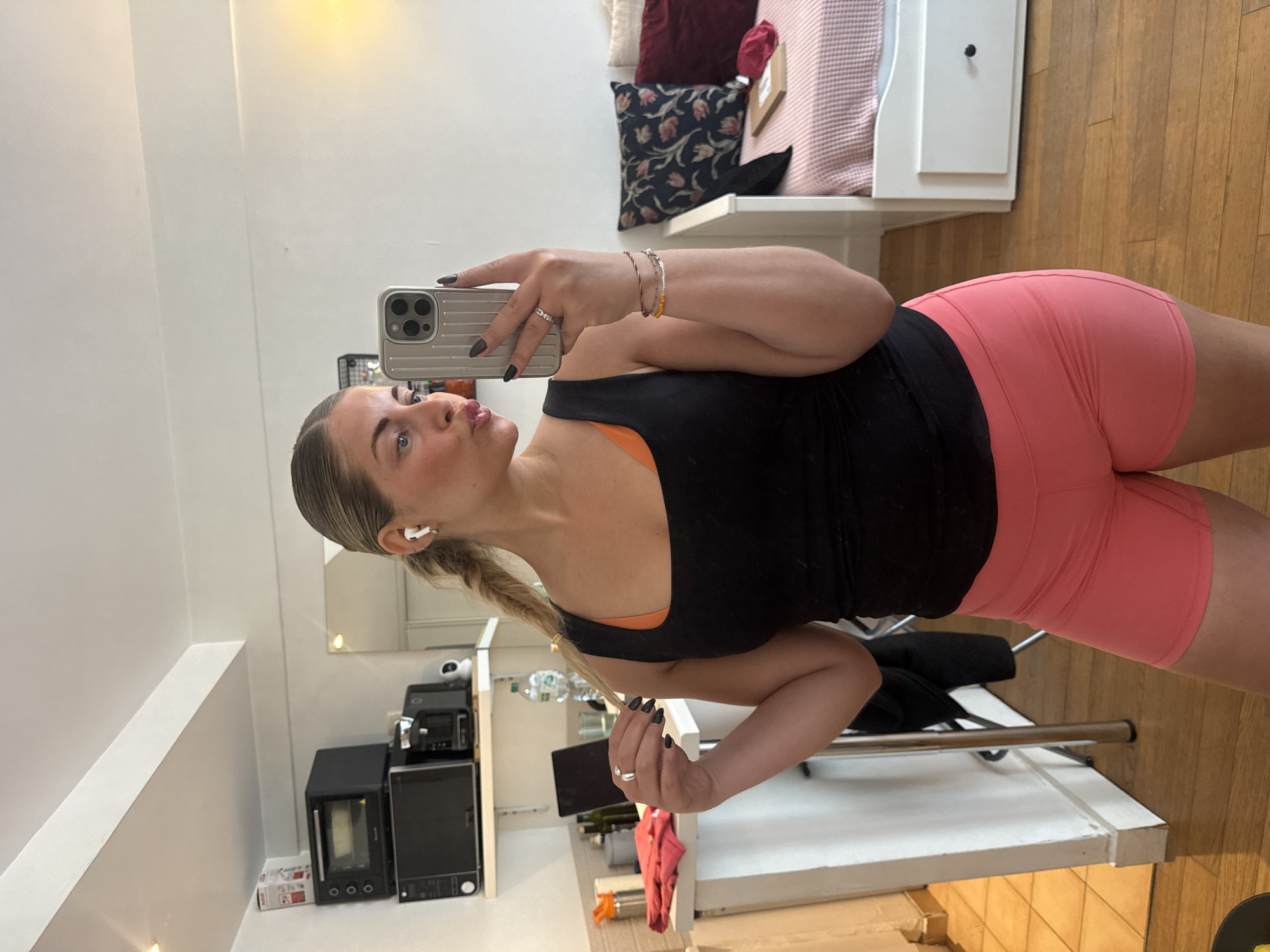 A person is standing indoors, holding a smartphone in their right hand to take a mirror selfie. The person has blonde hair tied back in a braid, and is wearing black tank top with orange straps and pink shorts. In the background, there is furniture including a white cabinet with pillows, kitchen appliances such as a microwave, and a counter with items on it. The location appears to be inside a kitchen or living space and not specifically identifiable as Paris, France based on the visual information provided.