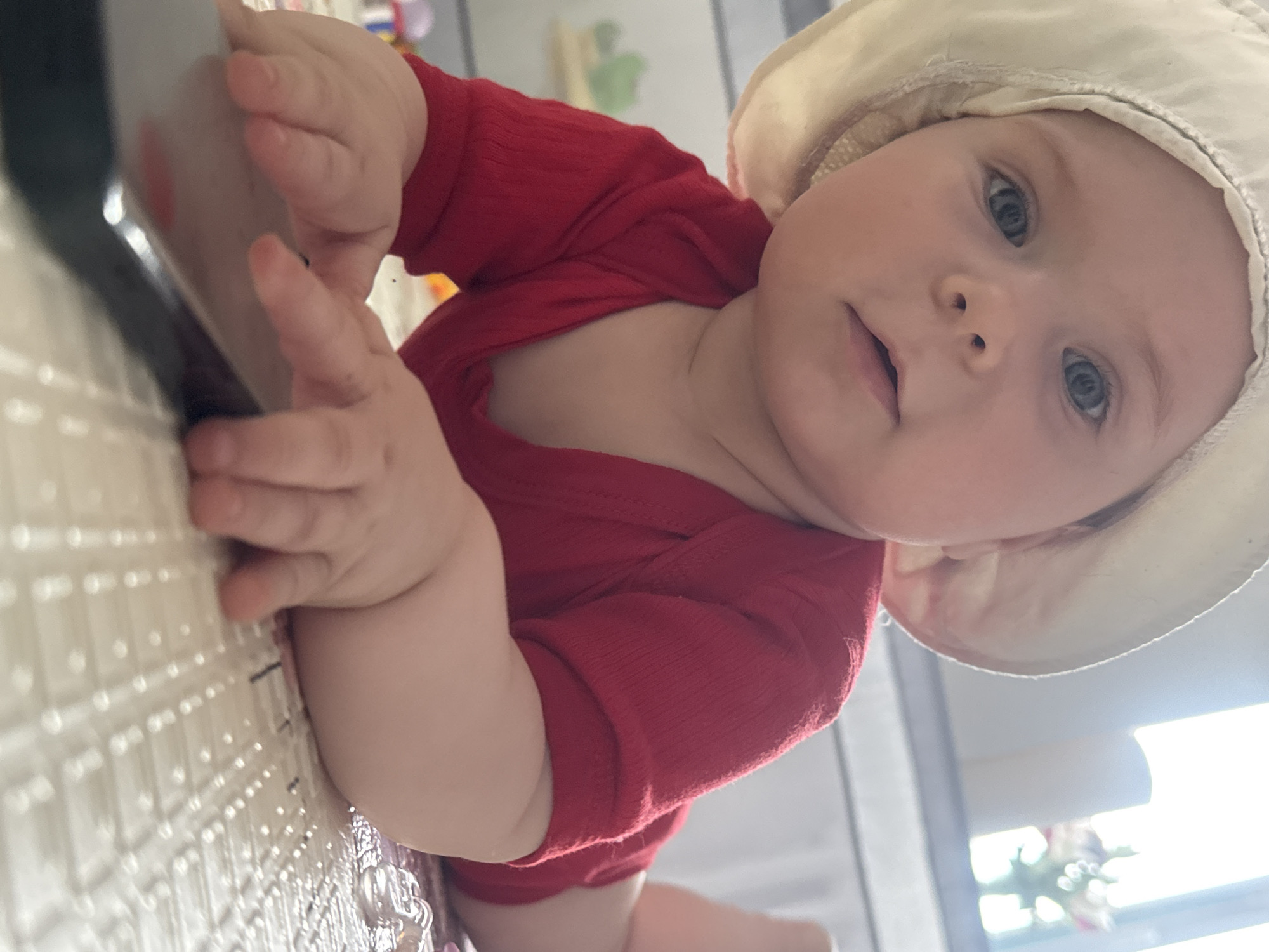 A baby wearing a red long-sleeved shirt and a light-colored hat is lying on a textured surface. The baby is looking towards the camera and is touching a black object with its left hand. The location is not discernible from the image.