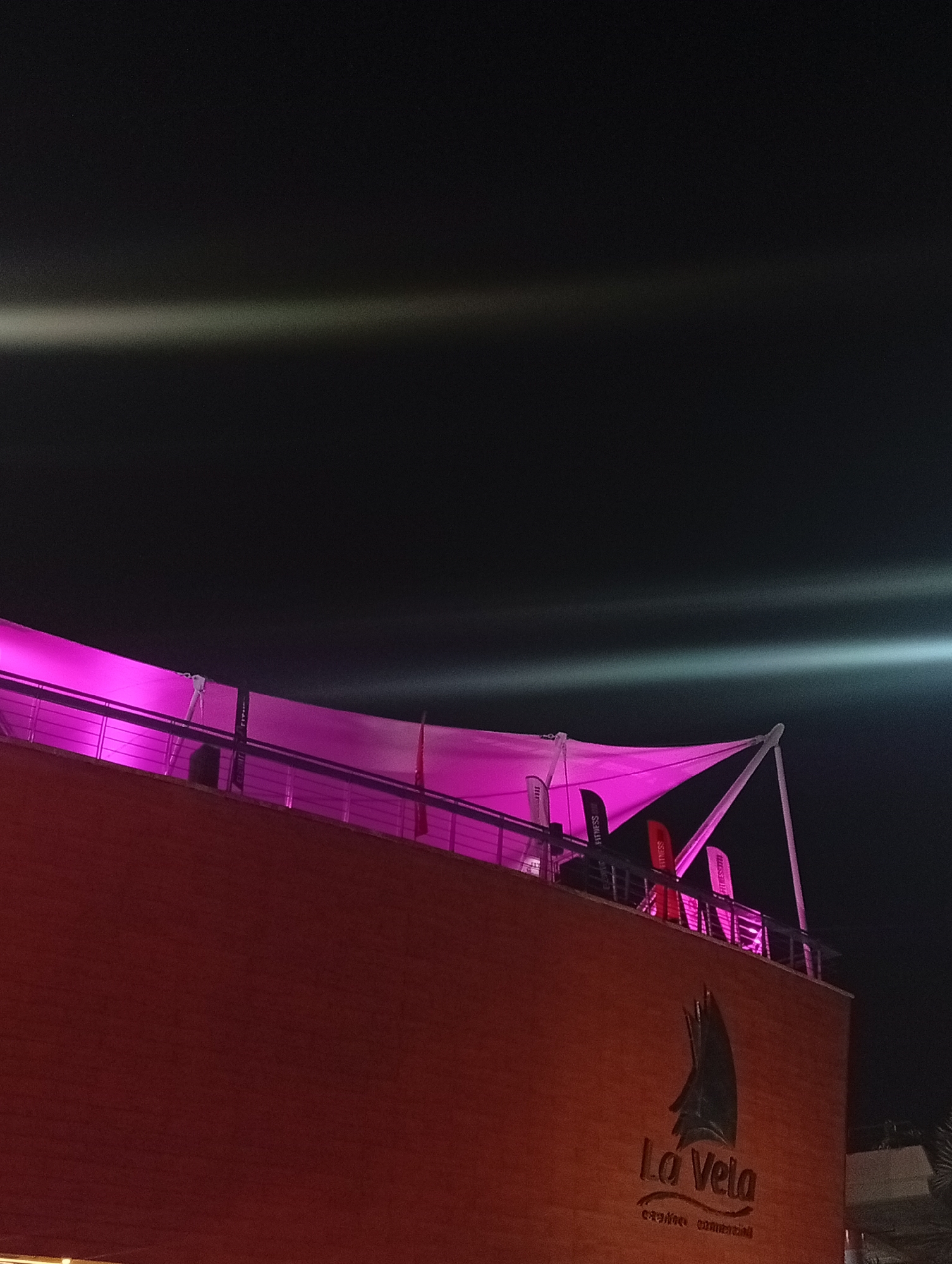 The image shows a low-angle, nighttime view of the "La Vela" business center in Porlamar, Venezuela. The building is lit with a pink hue. The roof has a canopy-like structure, also illuminated in pink, with supporting posts and railings. There are several "FITNESS SHOP" flags on the roof. The sky is dark with horizontal lens flares. The building is brick/brown color. There is some vegetation visible in the bottom right.