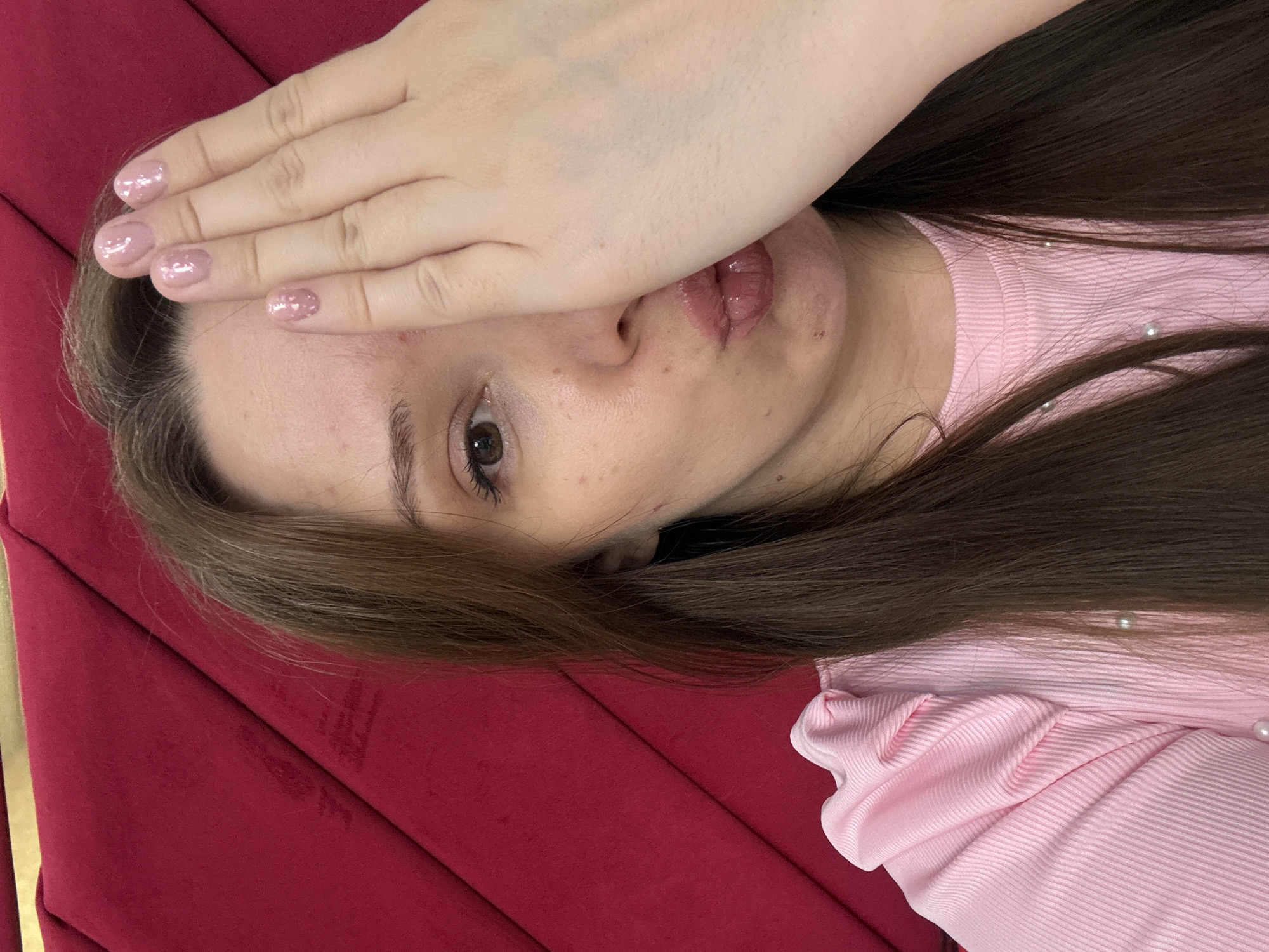 A close-up, slightly angled indoor selfie shows a young woman with long, dark brown hair covering her right side. She is wearing a pink, ribbed top with ruffled sleeves and pearl embellishments. Her left eye is visible, with brown irises and dark eyeliner. Her left hand is raised, covering the right side of her face, with short, manicured fingernails painted in a light pink, glittery polish. Her lips are pursed, and she is wearing a glossy pink lipstick. The background is a textured, dark red surface. The image appears to be taken in a residential setting, likely a home in Засечное, Russia.