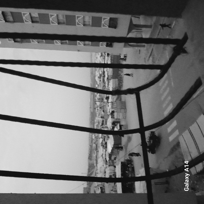 Black and white image of a cityscape viewed through a window's bars. The bars are curved and thick, obscuring some of the view. A multi-story building with repeating window patterns stands to the left. Below, the cityscape includes pedestrian walkways, roads with painted crosswalks, and small structures resembling shacks. Several small figures can be seen walking or standing in the distance. The text "Galaxy A14" is visible in the lower right corner. The perspective is slightly tilted or rotated.
