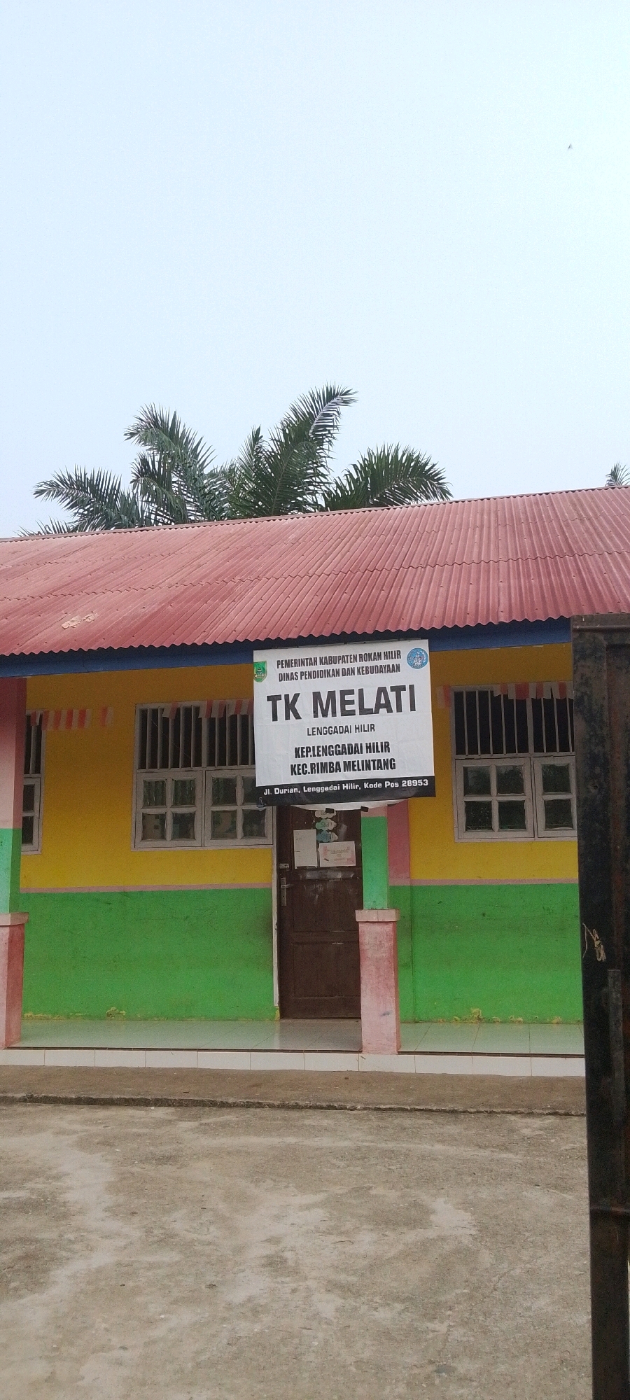 The image depicts the exterior of a school building, identified as TK Melati, located in Lenggadai Hilir, Rokan Hilir, Indonesia. The building has a red corrugated metal roof, a yellow upper wall, and a green lower wall. There are barred windows on both sides of a brown wooden door, which is centrally located on the building's facade. Above the door, a white sign displays the school's name, "TK MELATI," along with its address and administrative details: "PEMERINTAH KABUPATEN ROKAN HILIR DINAS PENDIDIKAN DAN KEBUDAYAAN," "LENGGADAI HILIR," "KEP.LENGGADAI HILIR," "KEC.RIMBA MELINTANG," and "Jl. Durian, Lenggadai Hilir, Kode Pos 28953." Palm trees are visible above the roofline. The foreground shows a paved ground surface, possibly a walkway or courtyard, with some concrete and a low retaining wall. A dark metal fence or gate is partially visible on the right side of the image. The sky is overcast.