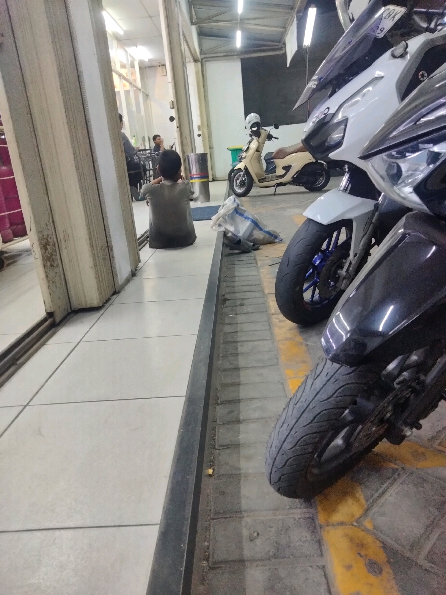 A medium-shot image captures a street scene in Pisangan Jaya, Indonesia. In the foreground, two motorcycles are parked side-by-side. The motorcycle on the right is black and partially obscures a white motorcycle to its left, which has blue accents on its wheels. A light-colored scooter with a white helmet is parked further back. In the mid-ground, a young boy wearing a grey t-shirt sits facing away from the camera, looking towards a table where two people are seated. The environment appears to be an outdoor or semi-covered area with a tiled floor and a metal grate along the edge of the pavement. The lighting suggests it may be evening or a dimly lit indoor space.