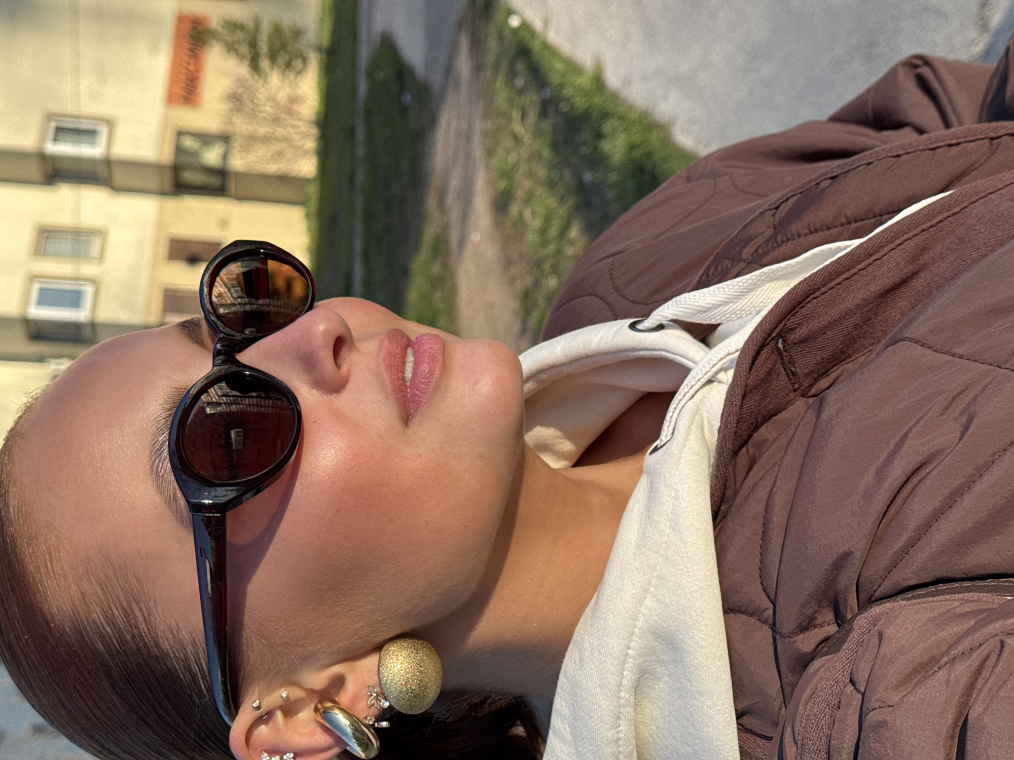 A medium shot of a woman wearing sunglasses, earrings, and a brown jacket with a white hoodie underneath. The lenses of the sunglasses reflect the image of a building, while the background consists of a paved pathway, grass, and other buildings. The location is Chaikovsky, Russia.