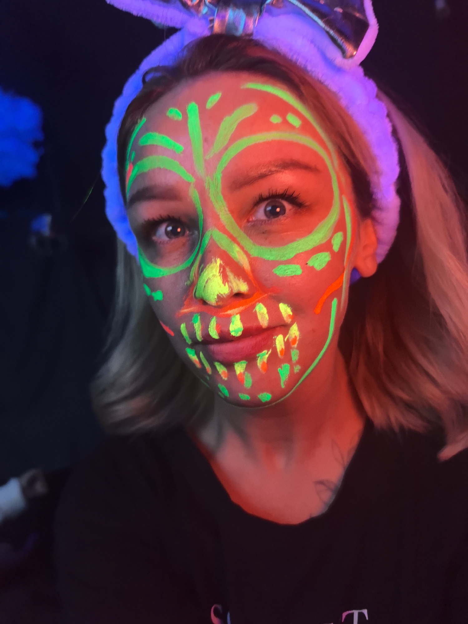 A person with blond hair is wearing a black t-shirt and a white headband with blue bunny ears. Their face is painted with neon green and orange designs that resemble a sugar skull. The nose is painted bright yellow. The background is dark. The scene is illuminated with red and blue lights. The location and city of Noviy, Russia cannot be determined from the image.