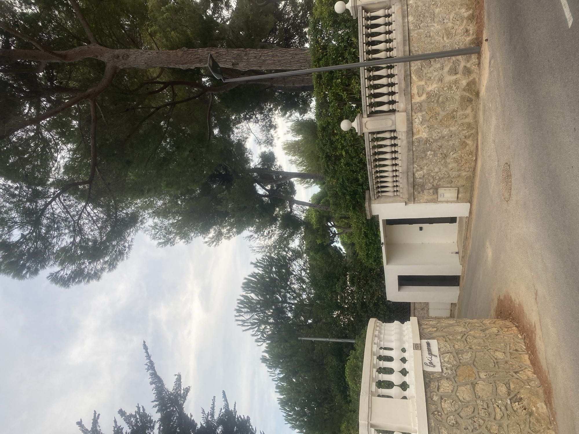 An exterior view captures a stone wall with a white railing and pillars leading to a modern white entrance. The wall features a sign with the inscription "Lociguanne 1325." Beyond the entrance, a paved driveway curves out of frame, flanked by dense green foliage and tall trees. A lamppost stands near the wall. The scene is set in Antibes, France, under a cloudy sky.