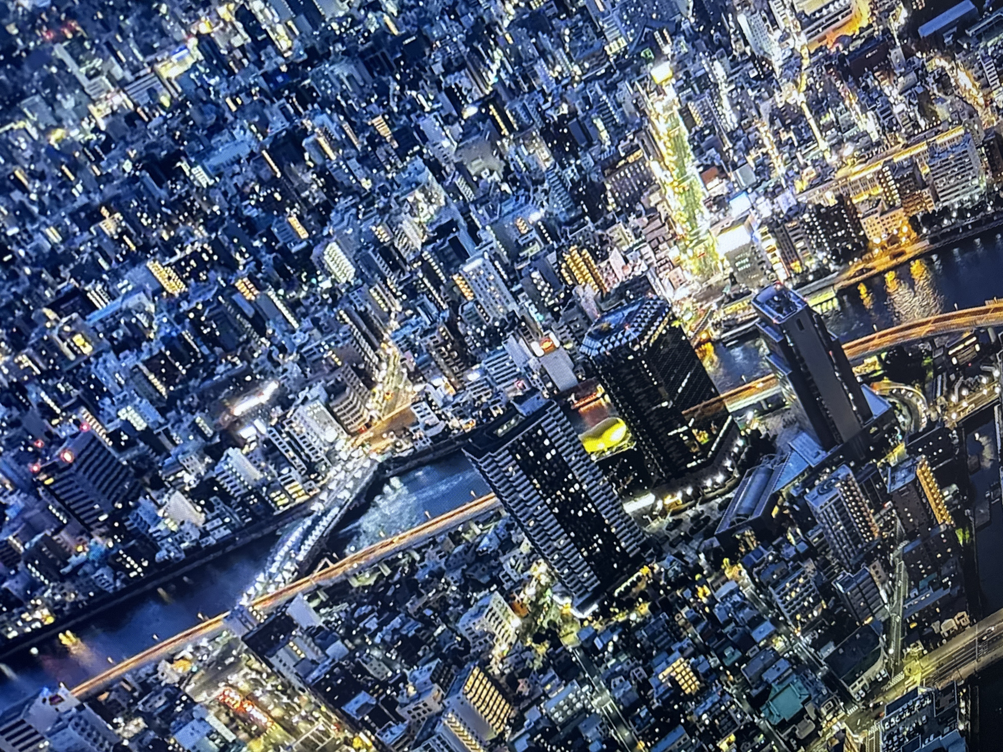 The media file presents an aerial, tilted view of a densely populated city at night, illuminated by a myriad of artificial lights. Numerous buildings of varying heights and architectural styles form a sprawling urban landscape. A wide river or canal, dark yet reflecting the city lights, flows through the scene, crossed by multiple illuminated bridges. One particularly prominent tall, dark building stands near the river, featuring a distinctive bright yellow, amorphous or flame-like structure on its roof or adjacent to it.
The time of day is clearly night, with the sky appearing dark and the city glowing with light. The weather seems clear, allowing for sharp visibility of lights and reflections on the water. The city is characterized by a dense network of residential and commercial structures. The artificial lighting is a mix of warm yellows, bright whites, and cooler blues, creating a dynamic glow across the urban fabric. No discernible human activity or legible text is visible from this high vantage point.