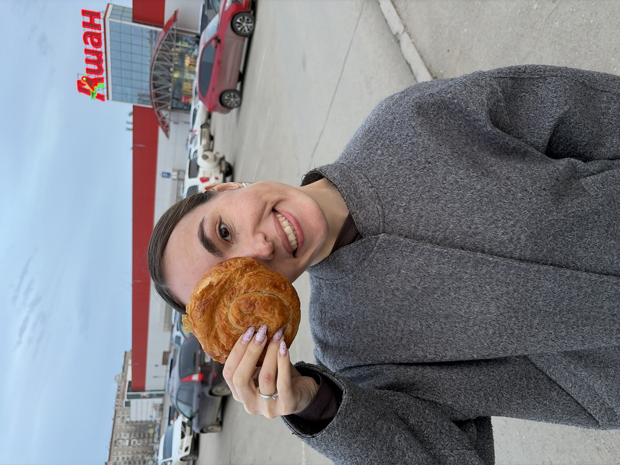 A woman in a gray coat smiles and holds a pastry. Behind her, cars fill a parking lot in front of a red and white building with "Auchan" written in red letters on the top. The sky is overcast. This is Izhevsk, Russia, as indicated by the Auchan store.