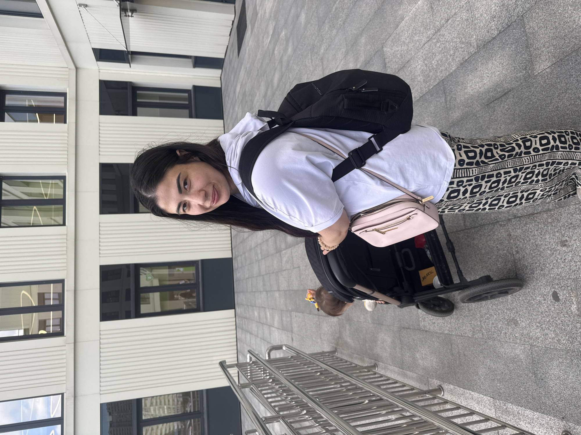 A young woman with long dark hair and a white hoodie stands outdoors, looking at the camera and smiling. She wears patterned pants and has a black backpack on her shoulders, with a light pink crossbody bag hanging from it. She is leaning on the handle of a black stroller, which is partially visible. A baby can be seen inside the stroller, wearing a yellow and blue toy on their head. The background features a modern building with horizontal white siding and dark-framed windows. The ground is paved with grey granite tiles. A metal railing is visible in the foreground. The scene appears to be set during the daytime, under overcast skies. The overall mood is casual and relaxed. There is no visible text in the image.