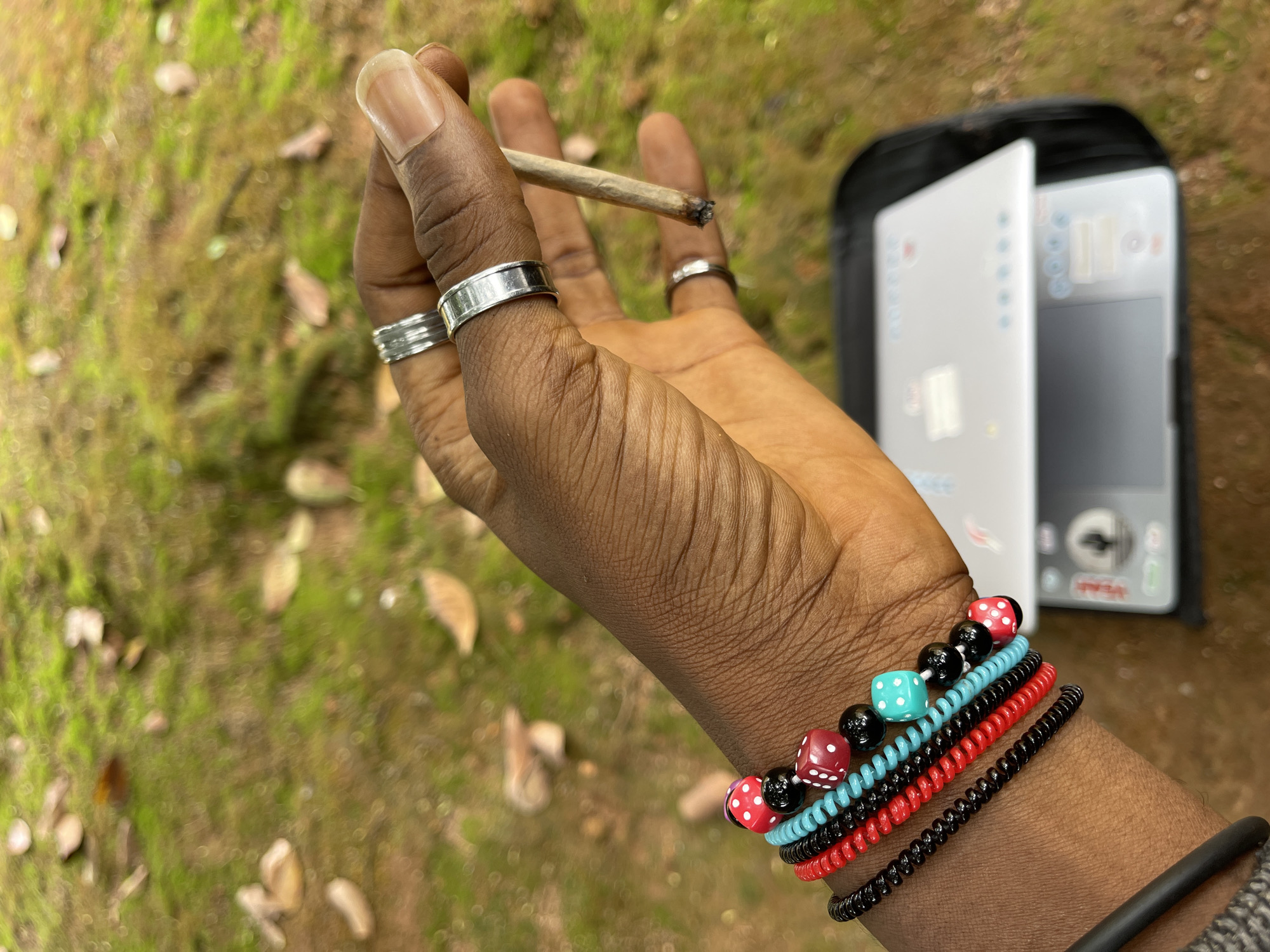 A hand holds a lit cigarette. The hand wears three silver rings on the fingers and several bracelets, including dice-themed bracelets, on the wrist. The background is blurred, but shows green vegetation and a laptop in a black case. Location information is not available in the image.