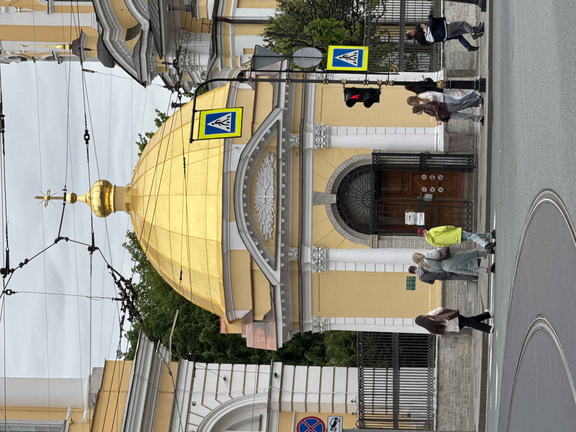 The image shows the Church of the Savior on Sennaya Square in Saint Petersburg, Russia, next to a crosswalk where people are crossing the street. The church has a golden dome and a yellow facade with white architectural details. Pedestrian crossing signs are visible. The sky is overcast. A tram line runs along the road.