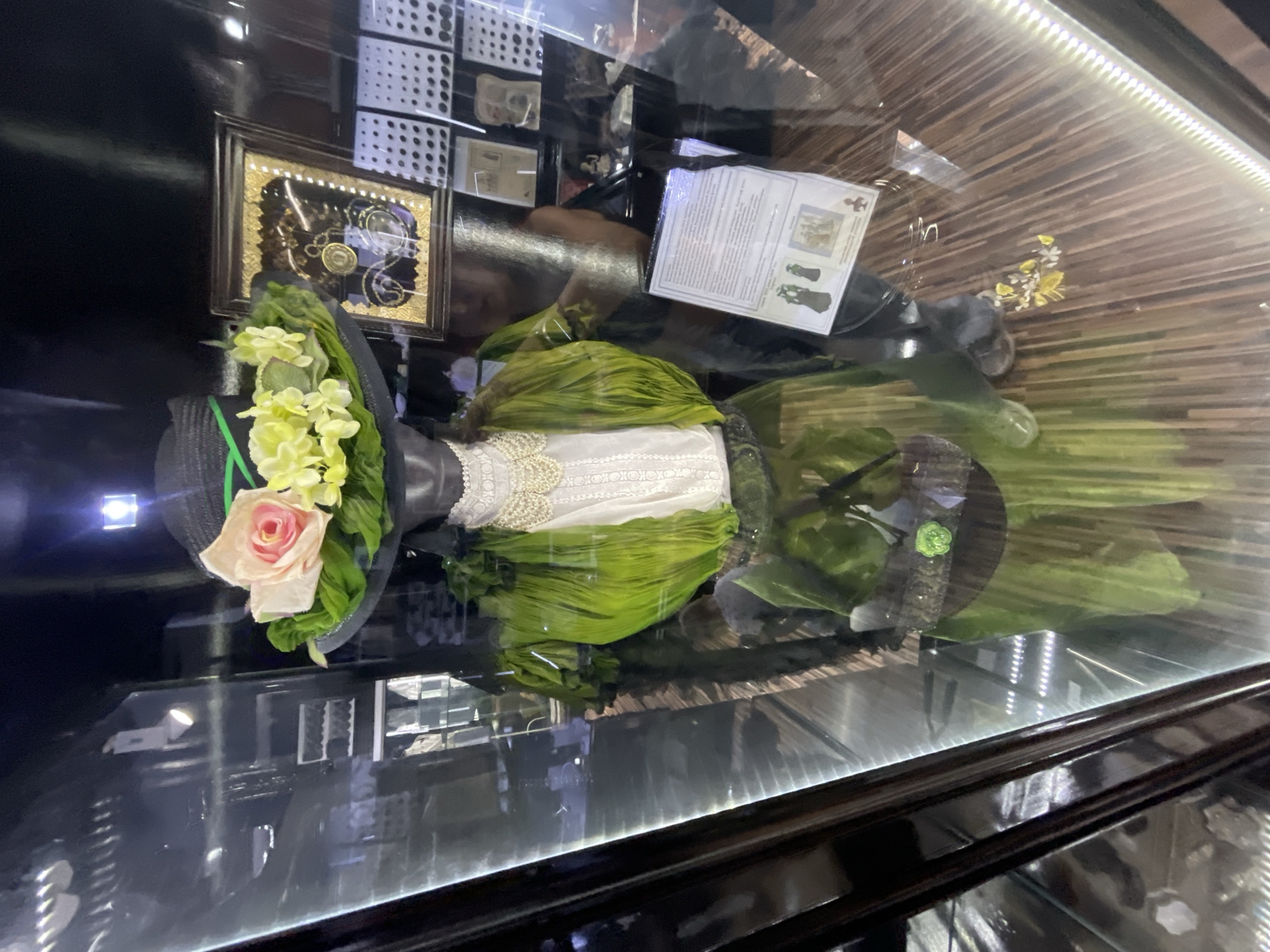 An interior shot shows a mannequin dressed in an antique green dress with layered fabric and a white lace collar. The mannequin wears a black hat adorned with a pink rose, yellow flowers, and green leaves. A document is displayed in front of the mannequin on a stand. The background reveals a museum display with framed items and shelves. The scene is lit with artificial lights, reflecting on the glass case.