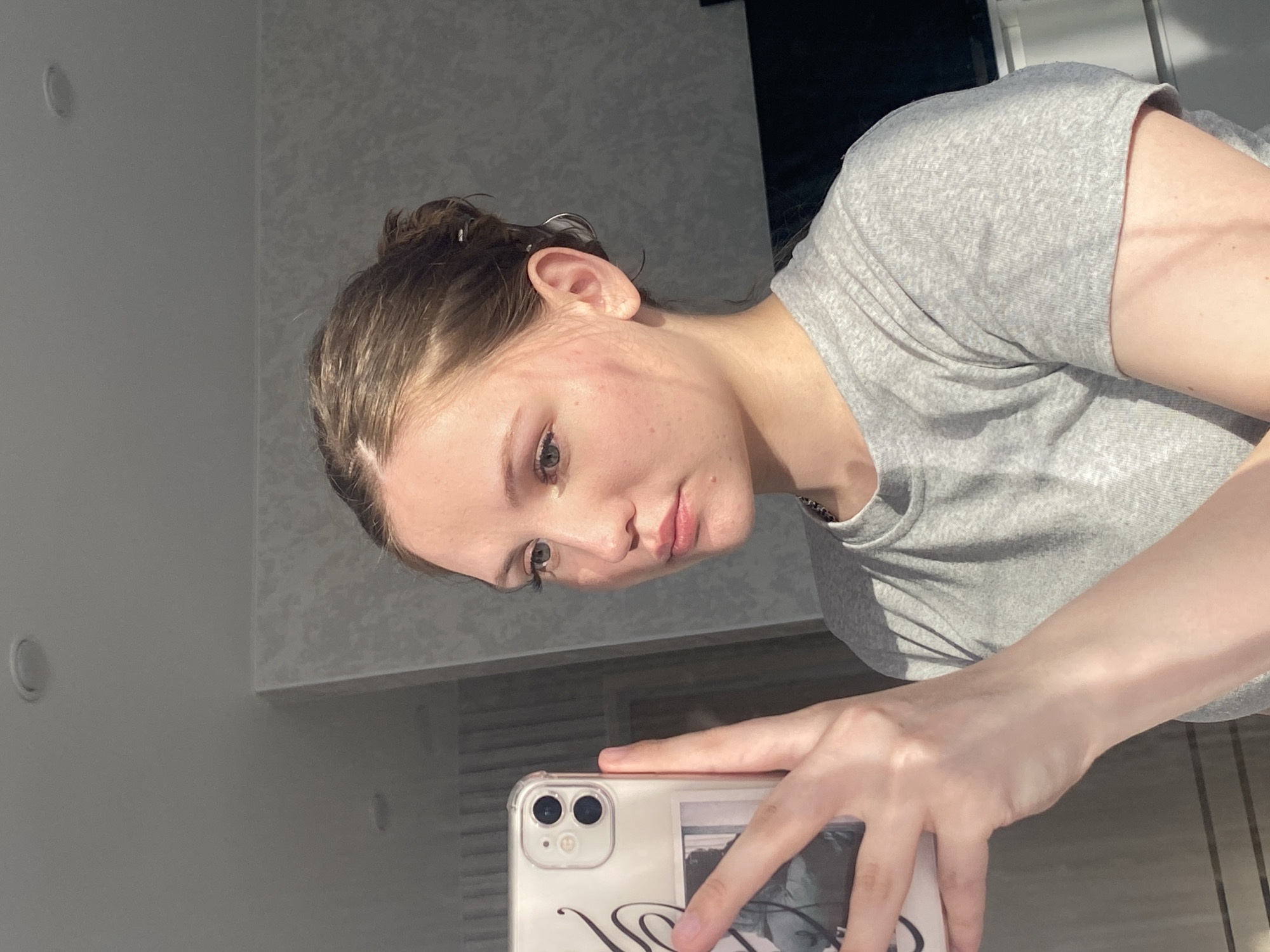 A young Caucasian woman is taking a selfie indoors. She is wearing a gray t-shirt. Her brown hair is pulled back. She is holding a white iPhone. The background appears to be a wall with light-colored paint. The location cannot be determined as Izhevsk, Russia based on the image.