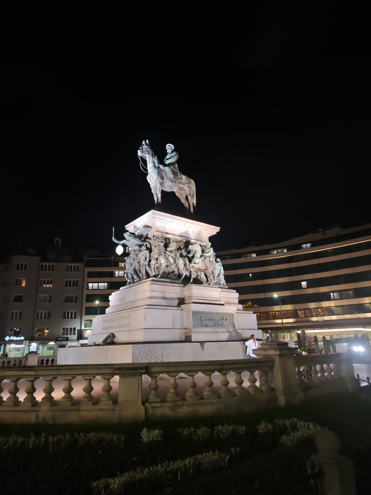 The image depicts the Monument to the Tsar Liberator in Sofia, Bulgaria, at night. A large equestrian statue of Tsar Alexander II stands atop a multi-tiered pedestal adorned with sculptural reliefs and figures. The monument is illuminated, contrasting with the dark night sky. Buildings with lit windows are visible in the background. A low stone wall with a balustrade surrounds a patch of vegetation at the base of the monument. Two individuals stand near the wall on the right side of the image. The overall scene is an urban landscape at night, focused on the historical monument.