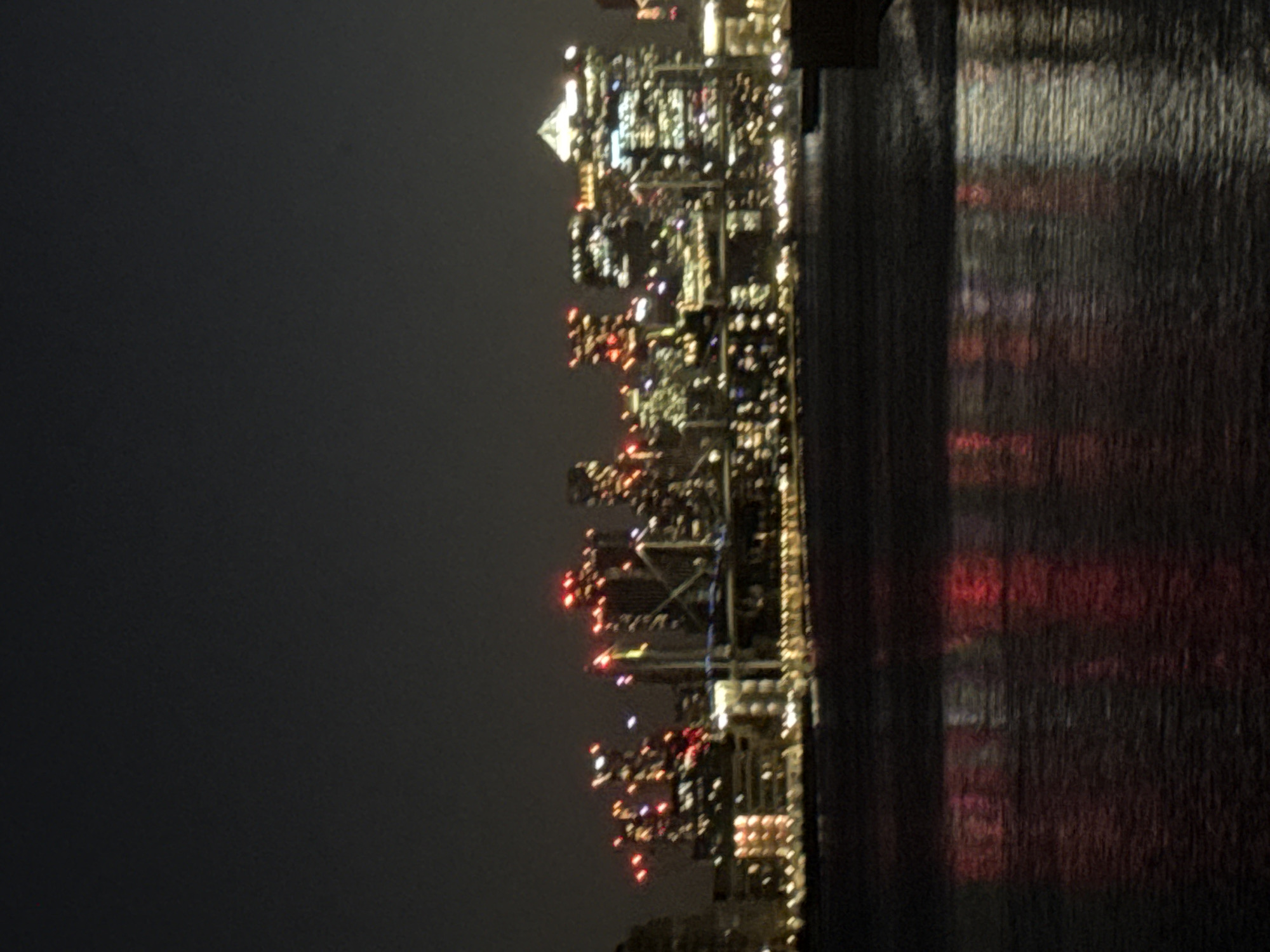The image shows a night scene of London, United Kingdom. The city skyline is visible with multiple buildings illuminated by numerous lights. The water reflects the lights from the buildings, creating a vibrant display. The darkness of the night contrasts with the bright city lights. The scene shows a mix of colors in the lights, and the water is textured and dark.