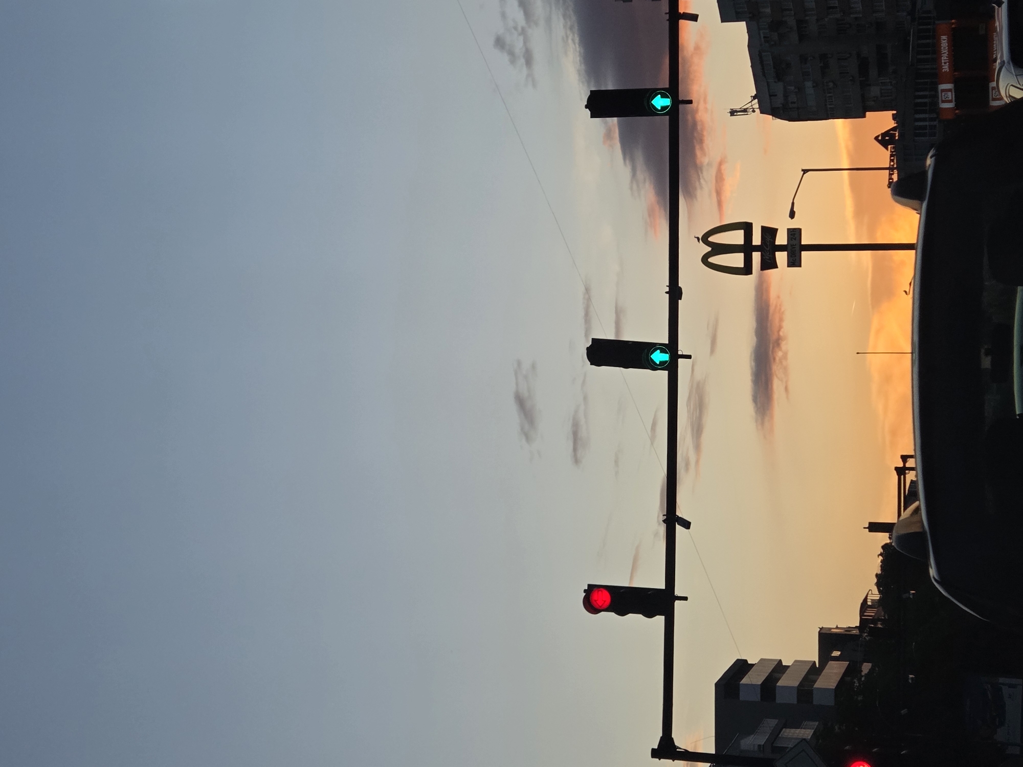 The image captures a street scene in Varna, Bulgaria, at dusk or dawn. The sky transitions from light blue to orange near the horizon. A traffic light pole stands prominently in the foreground. The topmost traffic light shows a green arrow, the middle shows a green arrow, and the bottommost traffic light shows red. In the background, there is a McDonald's sign, identifiable by the golden arches. Buildings, including an apartment block with visible balconies, are silhouetted against the sky. A street lamp is visible. Part of a car is in the right foreground. The McDonald's sign has "McCafe" written in Cyrillic below the golden arches. Further to the right of the McDonald's sign is a cylindrical-shaped gas cylinder.