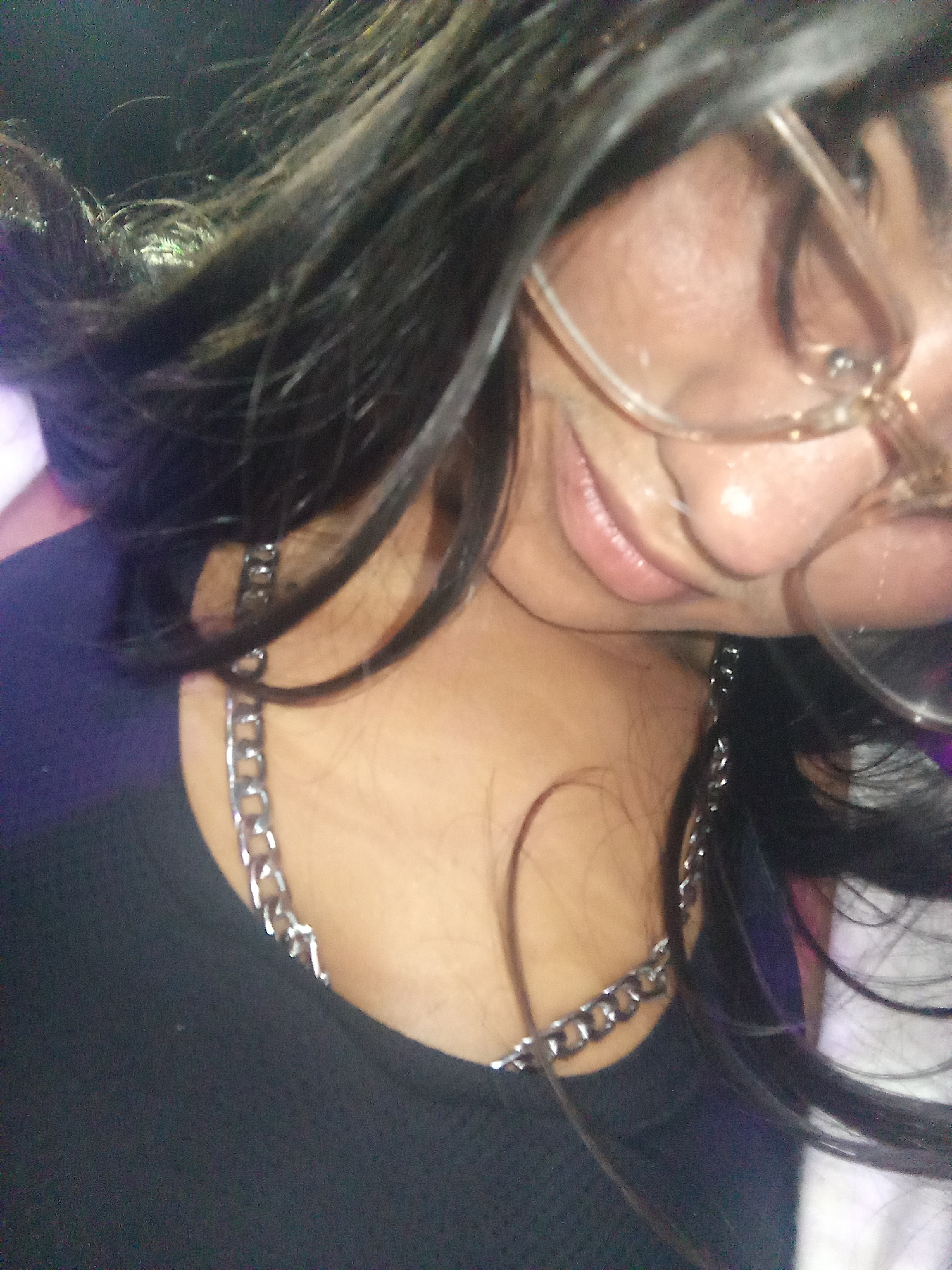 A close-up, angled shot depicts a person with dark hair, wearing eyeglasses with a light-colored frame, and a silver chain necklace. The person is wearing a dark-colored shirt. The lighting is dim, and the background is blurred. The photograph does not provide sufficient information to determine a specific location in Porlamar, Venezuela.