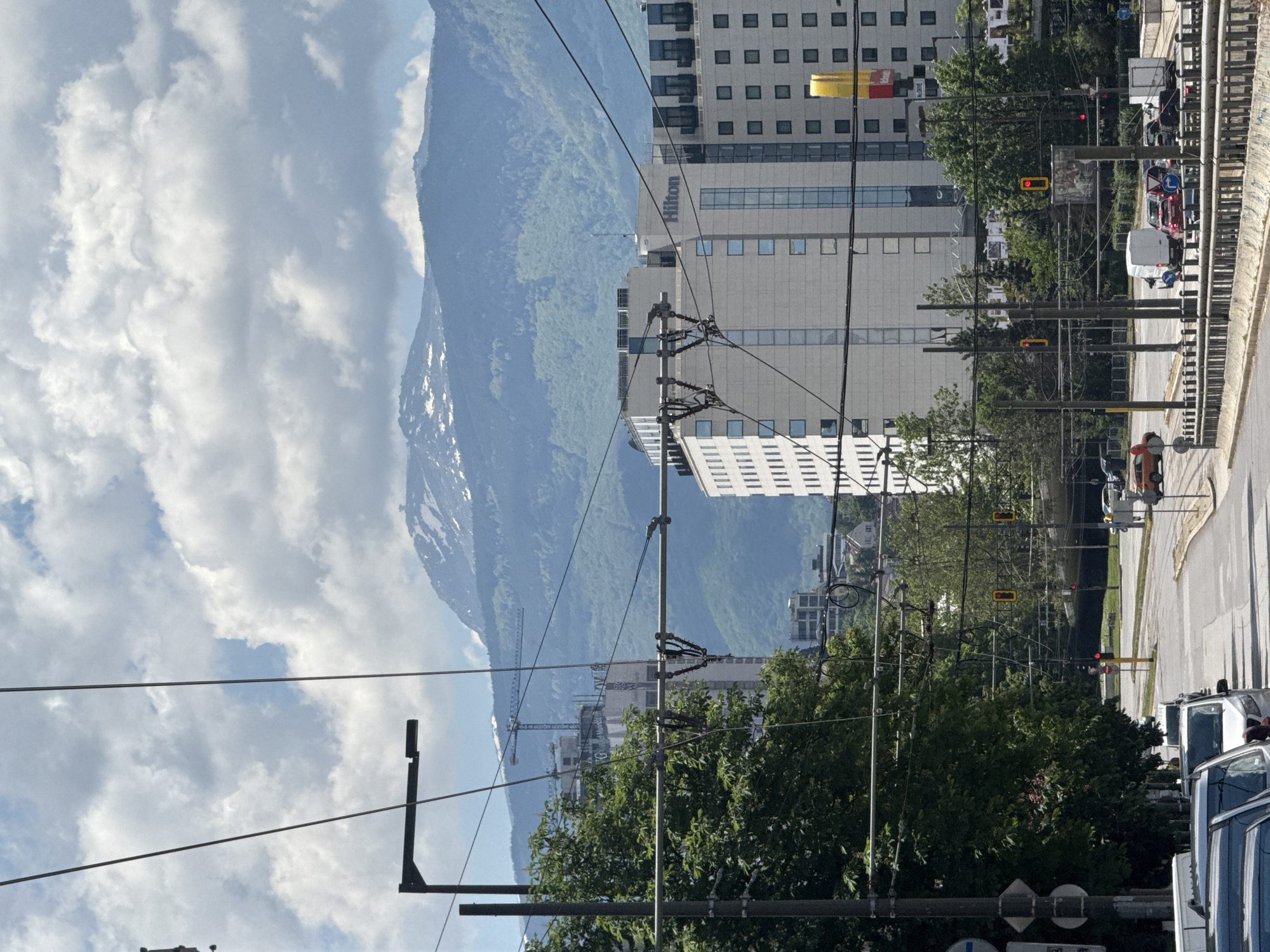 The image is a street scene in Sofia, Bulgaria, featuring a mountain range in the background and the Hilton Hotel in the midground. Overhead tram lines cross the street. A construction crane is visible behind trees. Traffic lights are present. The street has cars parked along the right side. The sky is cloudy.