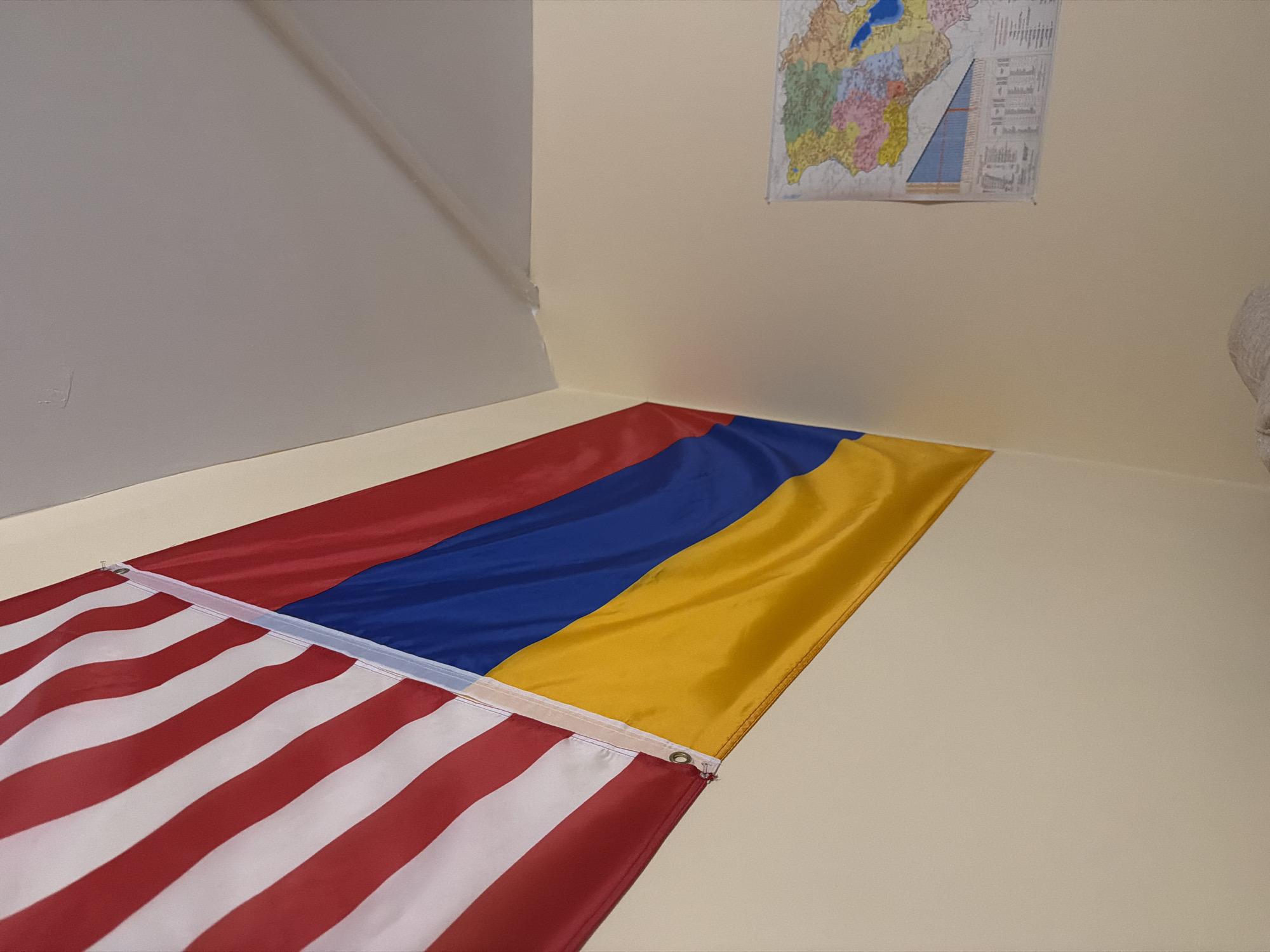 The image shows a combined flag of the United States of America and Armenia lying on the floor in the corner of a room. The American flag is at the bottom of the image, with its red and white stripes clearly visible. Above it is the Armenian flag, with its horizontal stripes of red, blue, and yellow. The flags are joined together by a white plastic strip.
In the background, there is a light-colored wall with a map hanging on it. The map appears to be of a geographical region with colorful sections. The room appears to be indoors and well-lit. There is no indication of the location being in Los Angeles, United States of America. There are no people in the image.