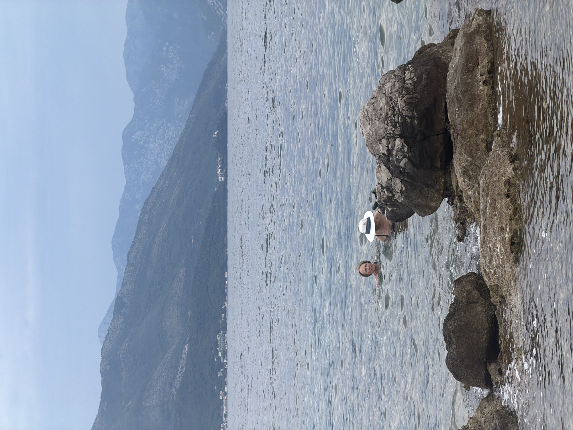 The scene depicts a body of water, likely the Adriatic Sea, in Gošići, Montenegro, as evidenced by the presence of mountains and coastal vegetation in the background. A woman wearing a white hat is partially submerged in the water, her head and shoulders visible. Rocks are prominent along the shoreline, some partially submerged. The water exhibits ripples and waves, indicating slight movement. The weather appears to be sunny, with a clear sky visible in the background. The overall environment is a coastal landscape with a combination of natural and possibly urban elements.