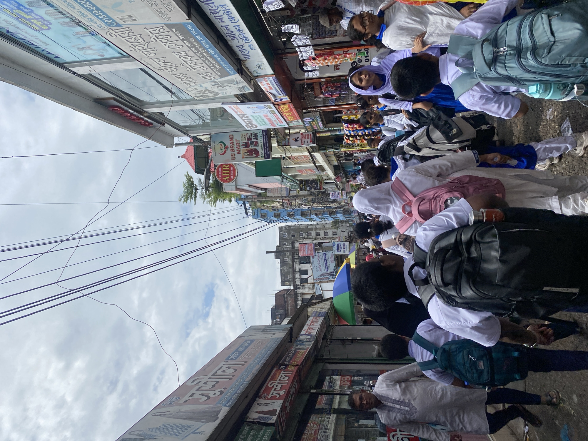 The image captures a bustling street scene in Dhaka, Bangladesh, likely a commercial area with shops lining both sides of a narrow street. A crowd of people, many of whom appear to be schoolchildren in uniform with backpacks, are walking along the street. The architecture suggests a dense urban environment with multiple-story buildings, some displaying signs in Bengali script. Utility wires are strung overhead, a common sight in Dhaka. The overall impression is of a busy market or commercial district during daytime hours.