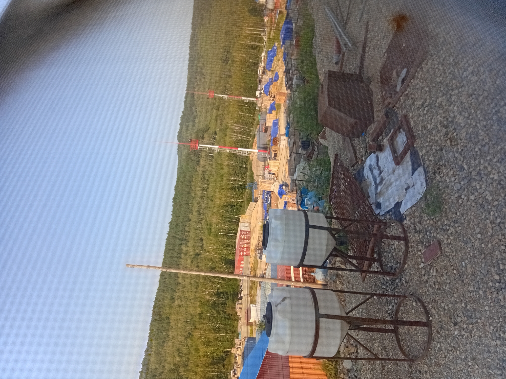 The image depicts a construction site with two large white water tanks in the foreground, supported by metal stands. Behind the tanks, a sprawling construction area is visible, featuring various structures, shipping containers, and numerous blue tarps covering materials. Two tall, white and red radio towers rise above the site. The background is dominated by a dense, green forest that extends up a hillside under a clear, pale blue sky. The ground is covered with gravel and scattered construction debris, including metal grates and a large, dark brown, angular object. A wooden pole stands vertically between the water tanks and the construction site. The scene appears to be located in or near Mtf Krasnaya Zvezda, Russia, given the natural landscape and industrial elements.