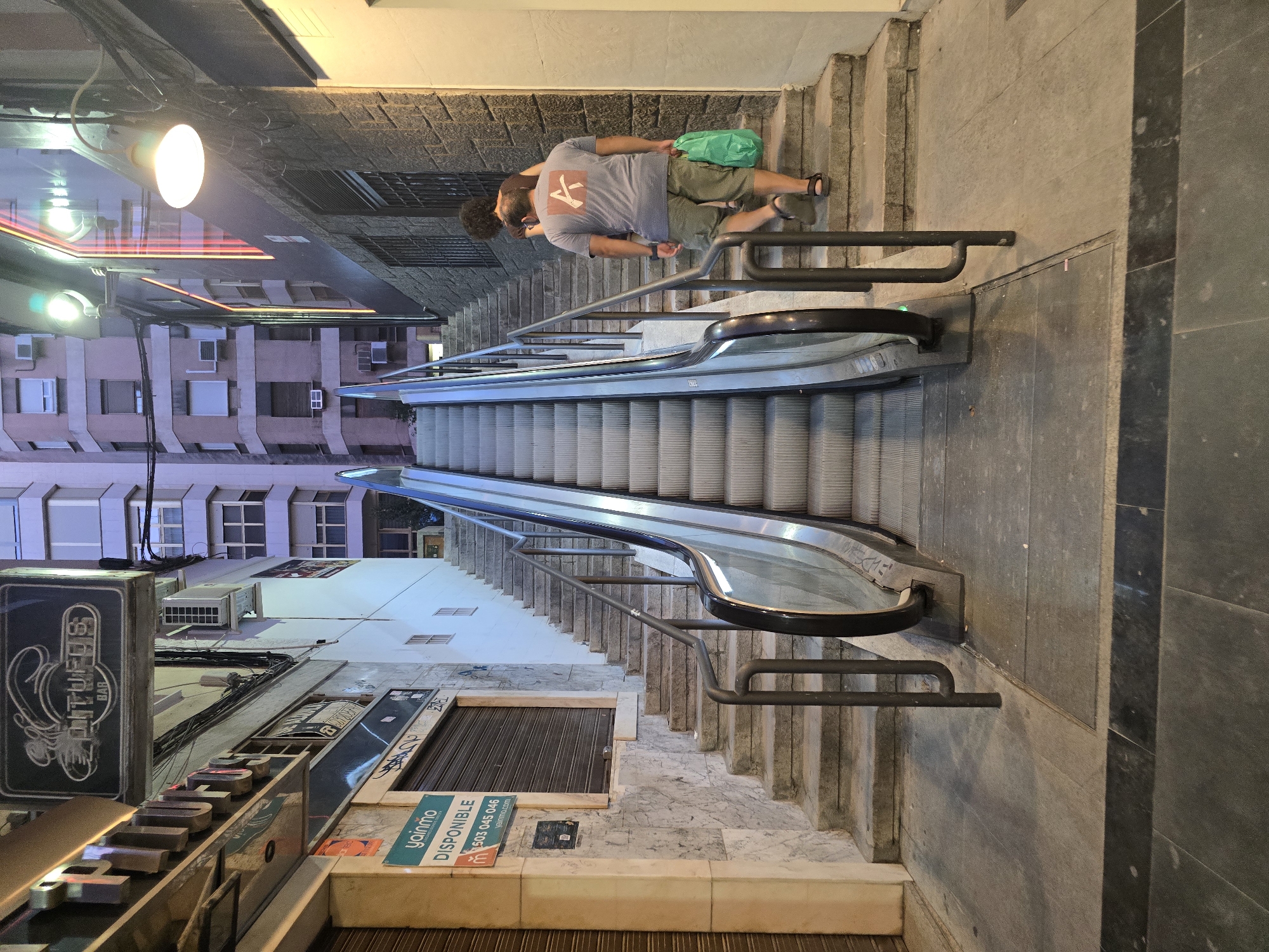 The image depicts an outdoor escalator and adjacent stairs in a narrow urban street, likely in the city of Jaén, Spain. The escalator is in the foreground and appears to be functional, with its moving steps visible. To the right of the escalator, a stone staircase ascends, and two individuals are walking up these stairs. The person in front is a woman with dark, curly hair, and the person behind her is a man wearing a grey t-shirt with a red logo on the back and khaki shorts. He is carrying a bright green plastic bag.
The buildings flanking the street are multi-story structures, with some featuring windows and air conditioning units. One building on the left has signage, including "YAINMO" and "DISPONIBLE," suggesting a business or service. A vintage-style sign with "LLITUFOS BAR" is also visible. The overall lighting suggests it is either dusk or night, with artificial lights illuminating the scene. The ground is paved with stone slabs, and some wear and tear are visible on the surfaces. The environment appears to be a commercial or residential area within a city.
